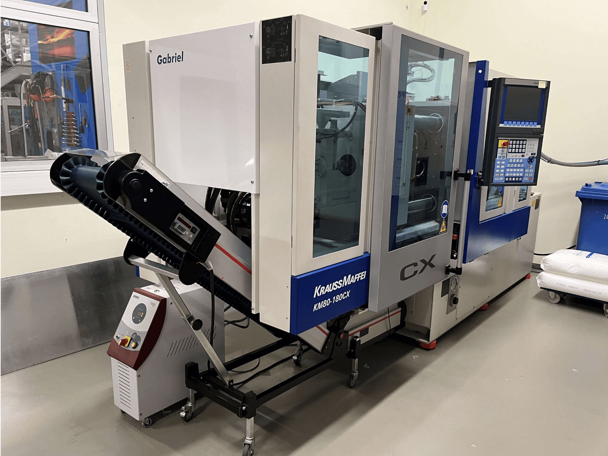 Máquina de moldagem por injeção hidráulica Krauss Maffei KM 80-180 CX, vista frontal, com painel de controlo e sistema de transporte.