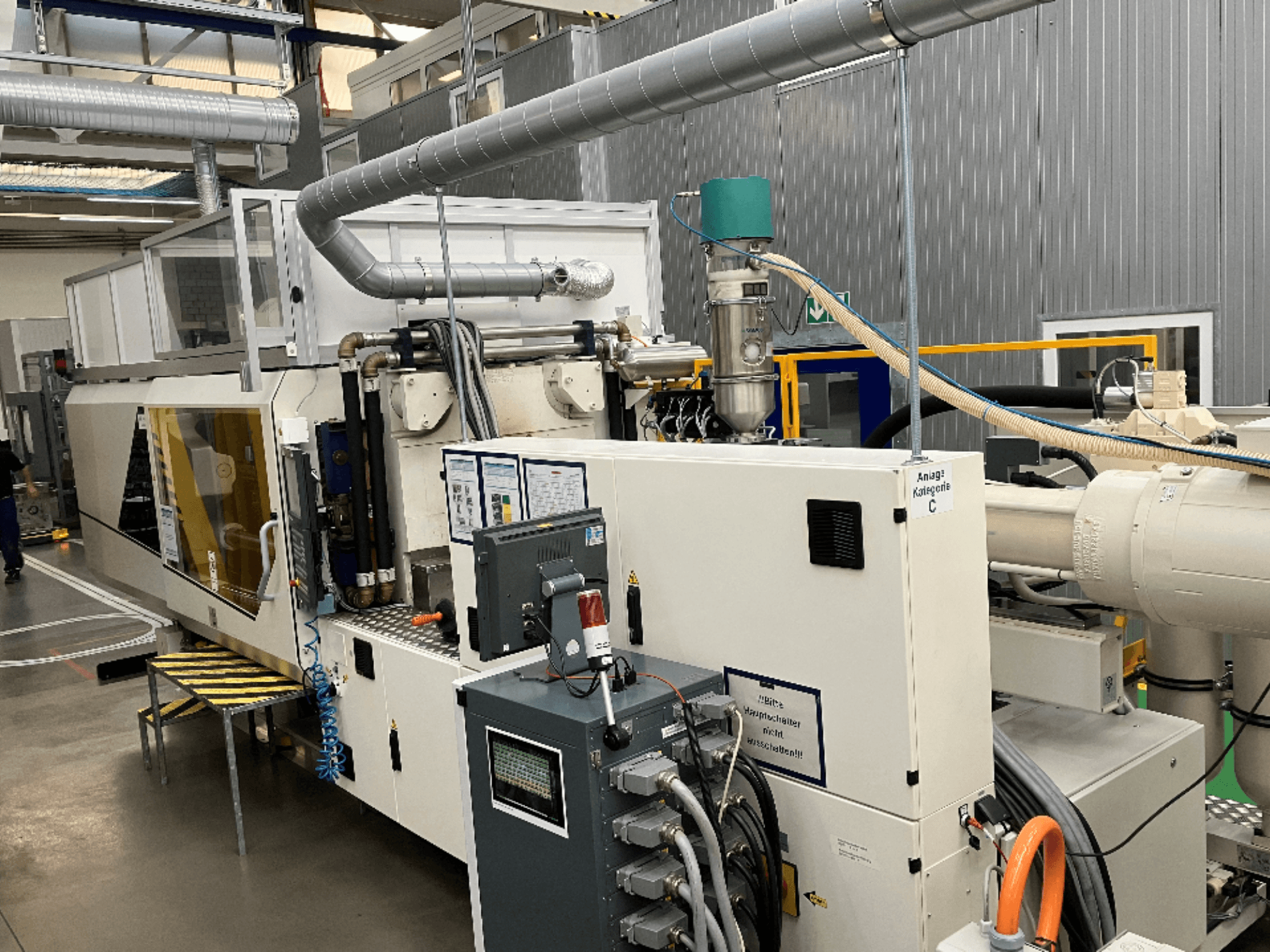 Máquina de moldagem por injeção, vista frontal, com etiqueta da marca, painel de controlo e mangueiras visíveis num ambiente de fábrica.