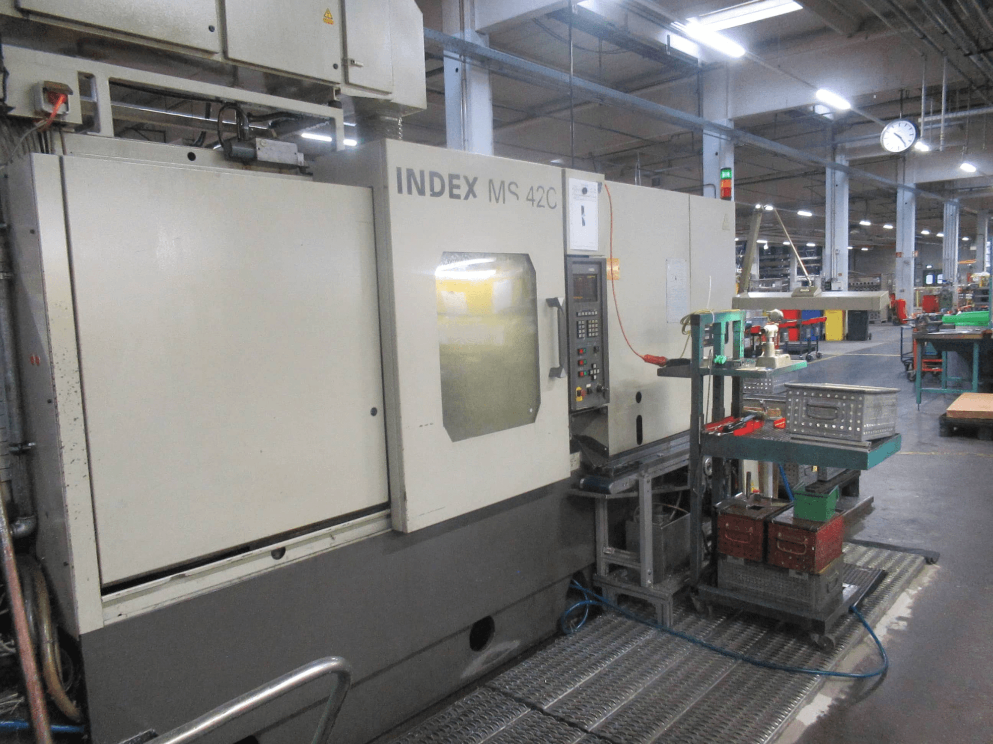 Máquina CNC INDEX MS 42C numa oficina, vista lateral mostrando o painel de controlo e o armazenamento de ferramentas num carrinho com rodas.