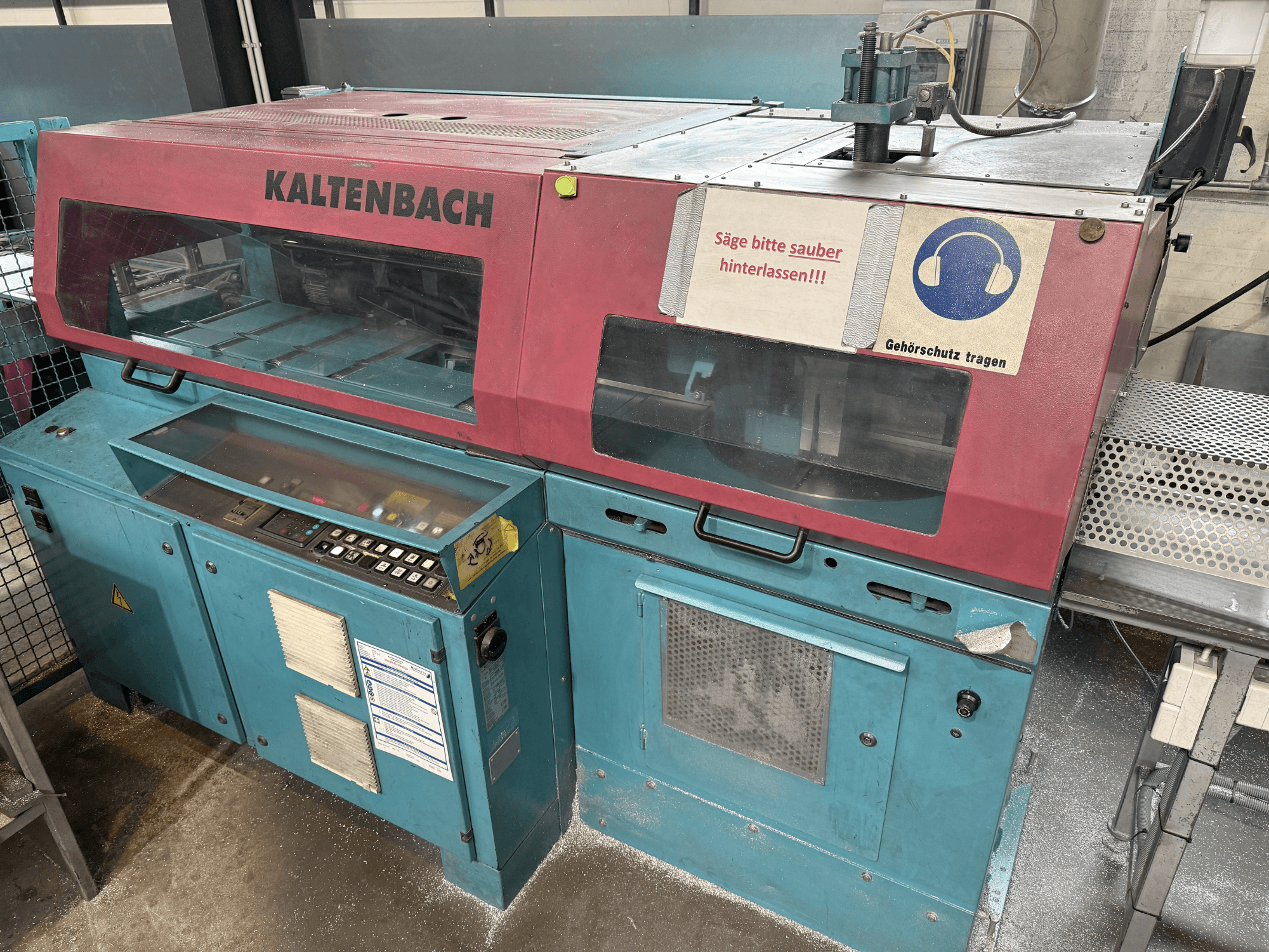 Máquina de corte industrial KALTENBACH, vista frontal, com painel de controlo e sinal de segurança para proteção auditiva.