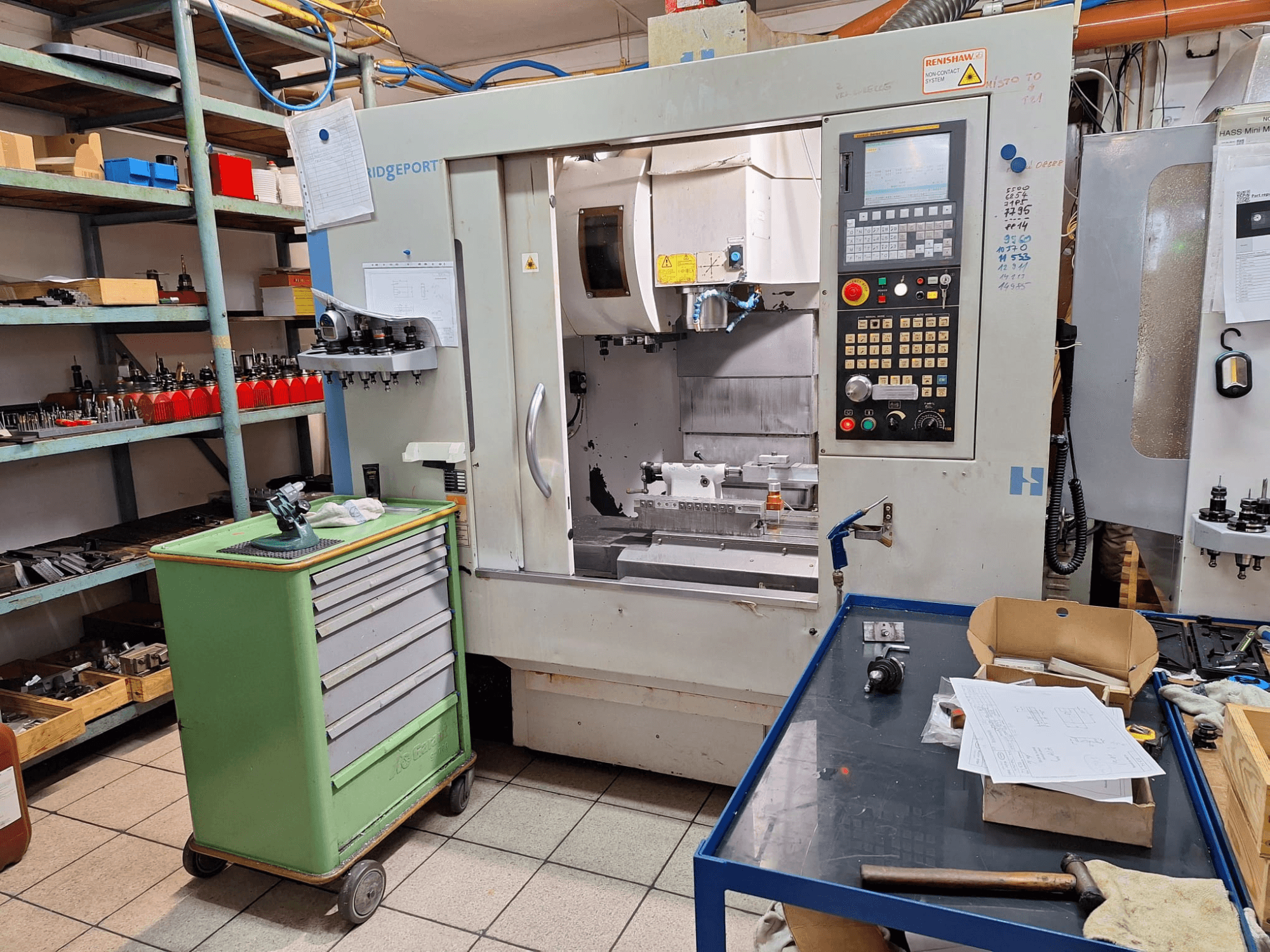 Vista frontal do centro de maquinagem vertical Hardinge Bridgeport GX 710, com painel de controlo, ferramentas e prateleiras, num ambiente de oficina.