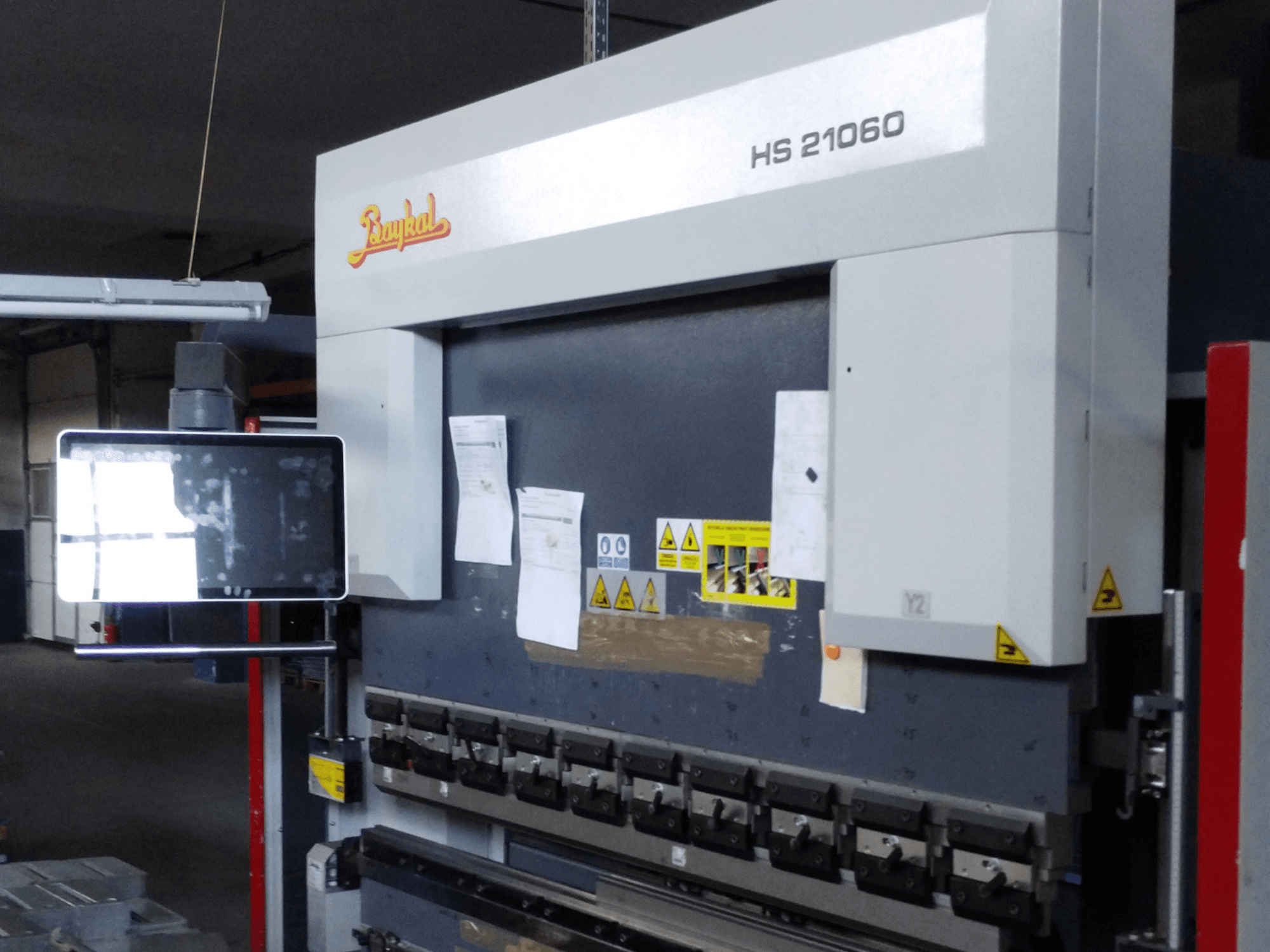 Vista frontal da prensa dobradeira Baykal APHS 21060, com ecrã de controlo, avisos de segurança e ferramentas para dobrar metal.