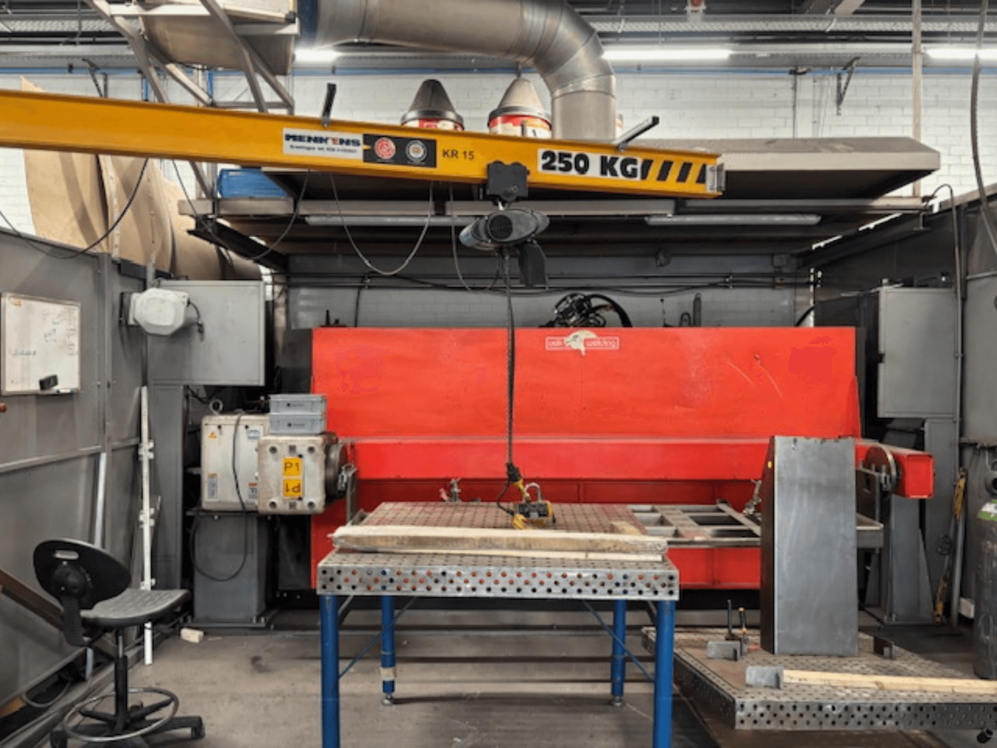 Oficina industrial com uma prensa hidráulica vermelha, uma ponte rolante com a etiqueta "KR 15" e uma mesa de trabalho em metal, perspetiva lateral.