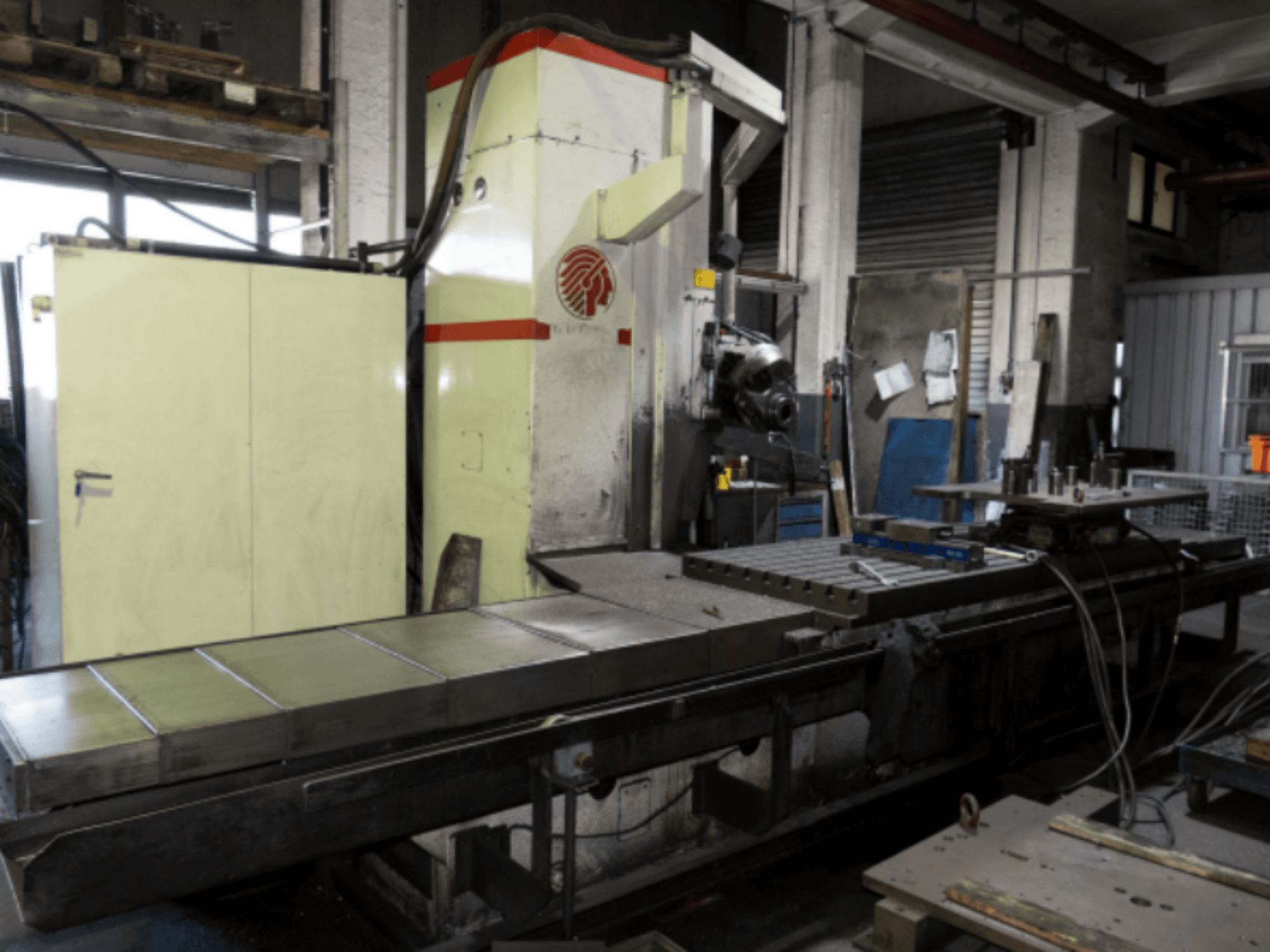 Fresadora industrial com uma mesa grande, vista lateral, mostrando controlos e acessórios numa oficina.