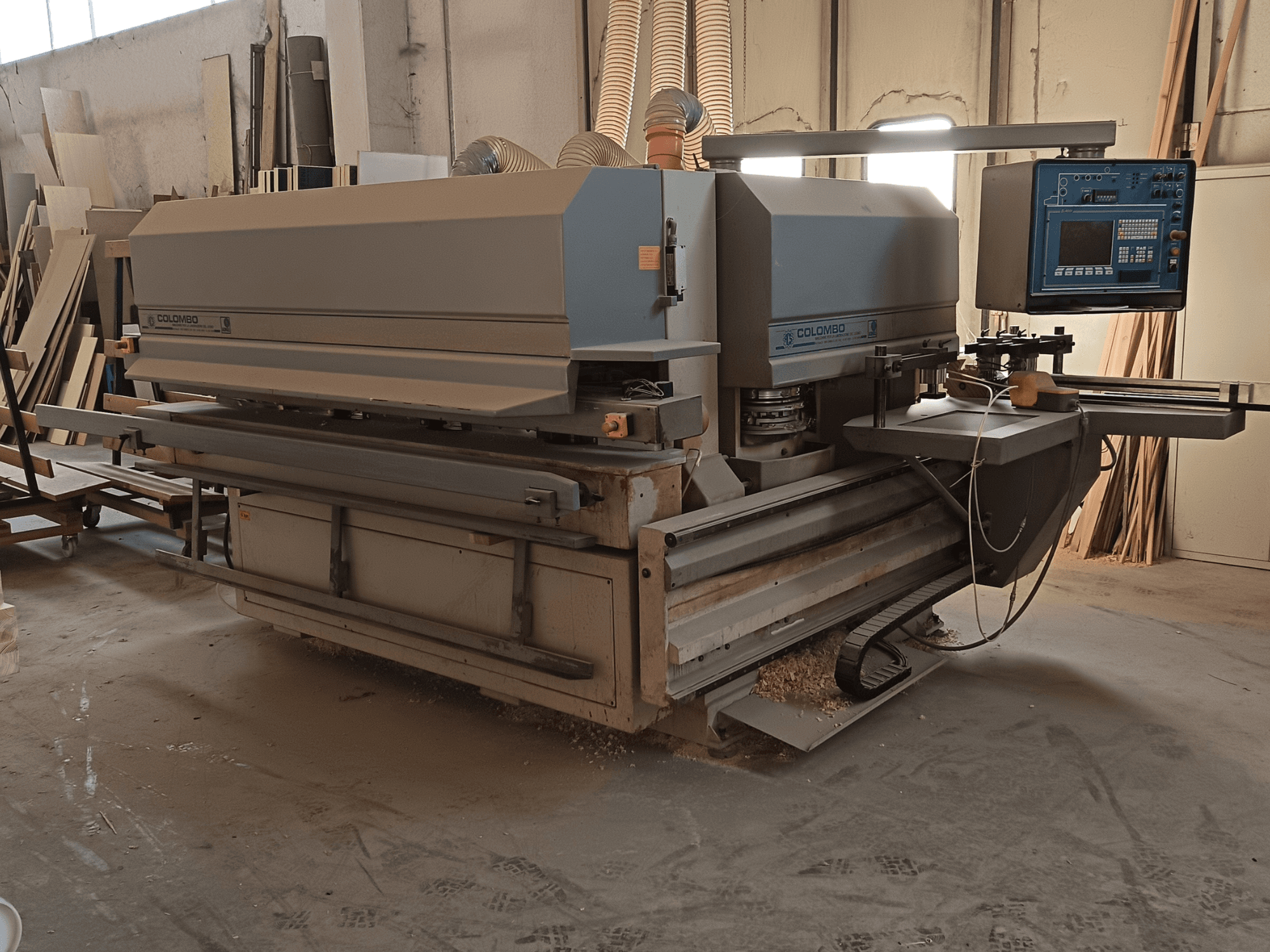 Máquina para trabalhar madeira Colombo, vista lateral, mostrando o painel de controlo, as lâminas de corte e a acumulação de pó de serra na oficina.