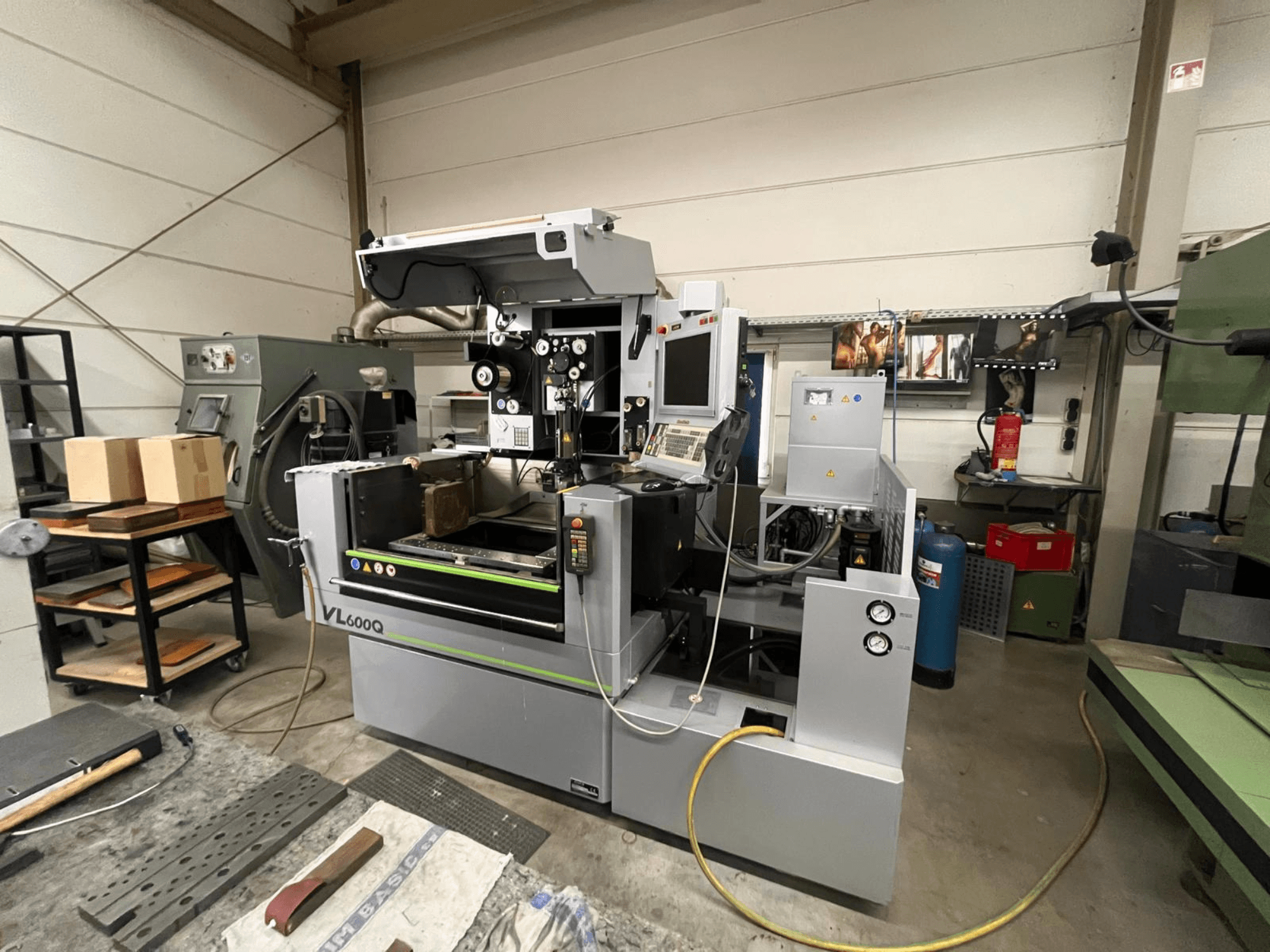 Máquina CNC VL600Q numa oficina, vista frontal, com painel de controlo, material colocado e várias ferramentas por perto.