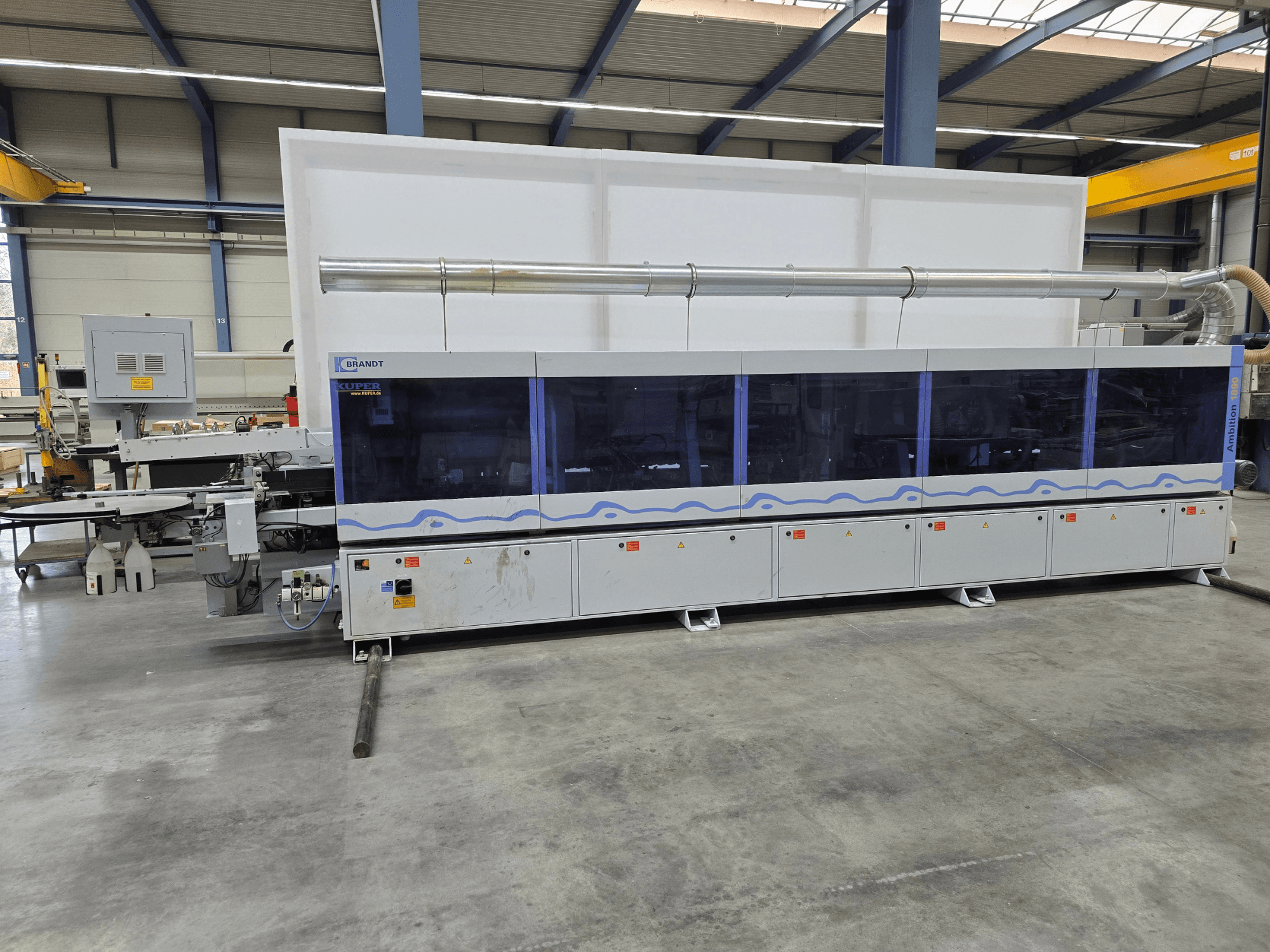 Máquina Brandt KUPER numa fábrica, vista frontal com painéis transparentes, com botões de controlo e avisos de segurança.