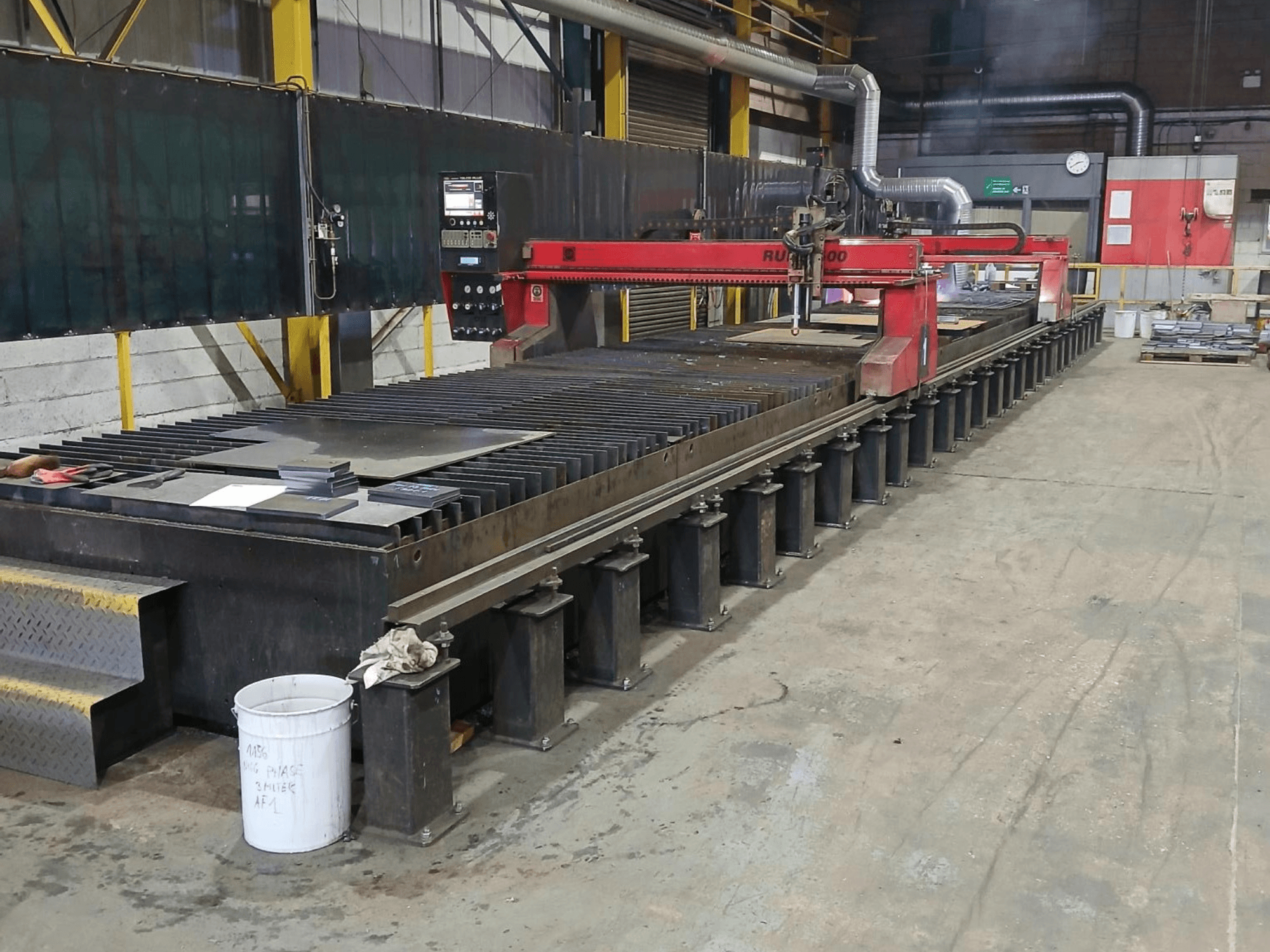 Máquina de corte RUIDA 300 vermelha, vista lateral, com chapa metálica sobre uma mesa de aço, situada num espaço de trabalho industrial.