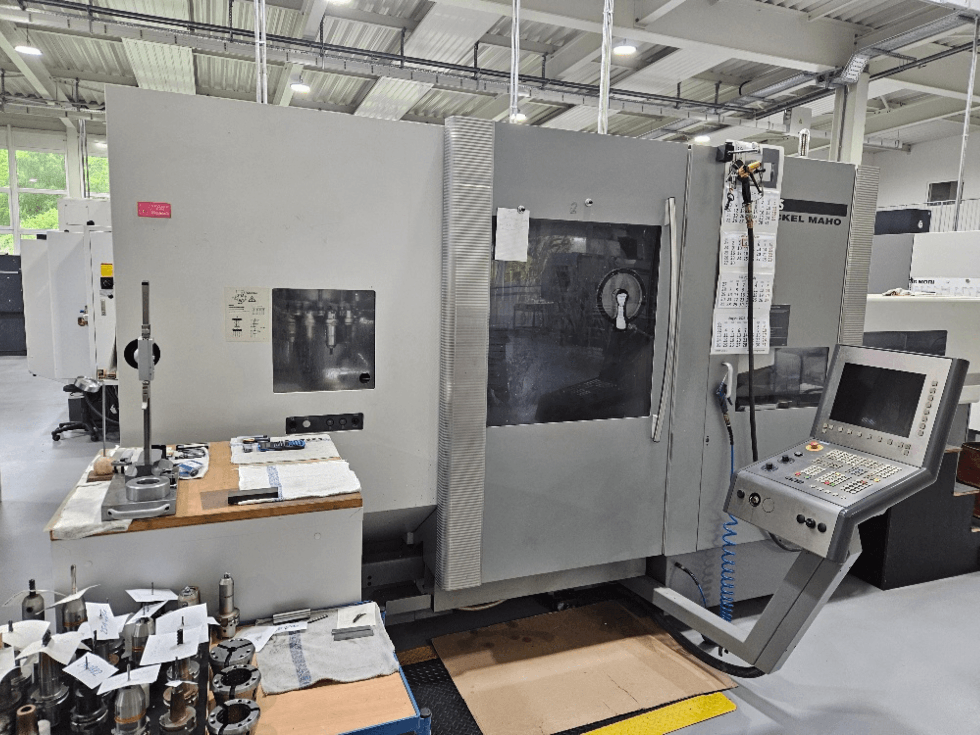 Vista frontal de uma máquina DMG Mori DMU 70 com painel de controlo, ferramentas numa mesa de madeira e várias peças de trabalho nas proximidades.