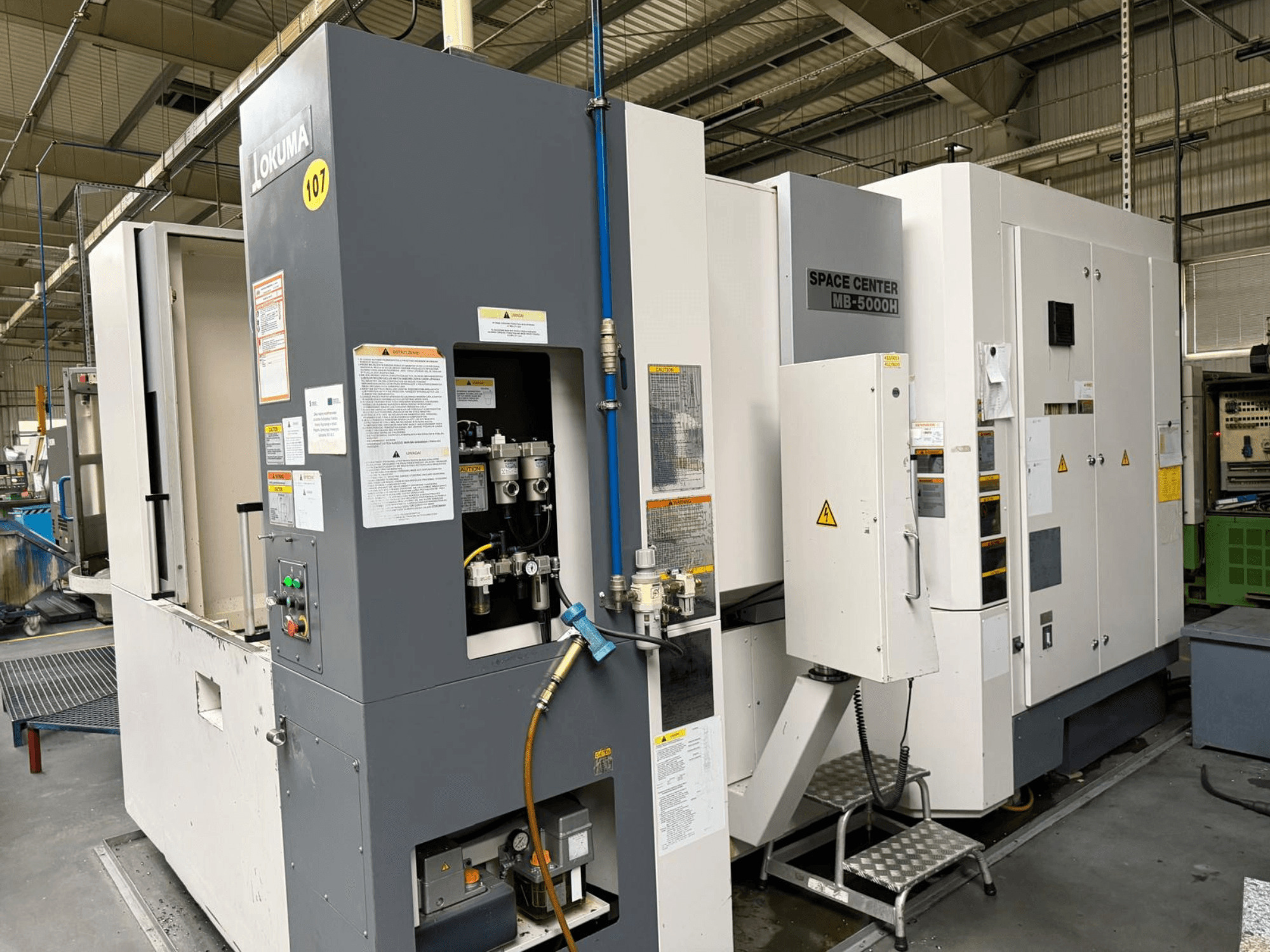 Centro de maquinação horizontal Okuma MB 5000H, vista lateral mostrando o painel de controlo e os componentes da máquina num ambiente de fábrica.