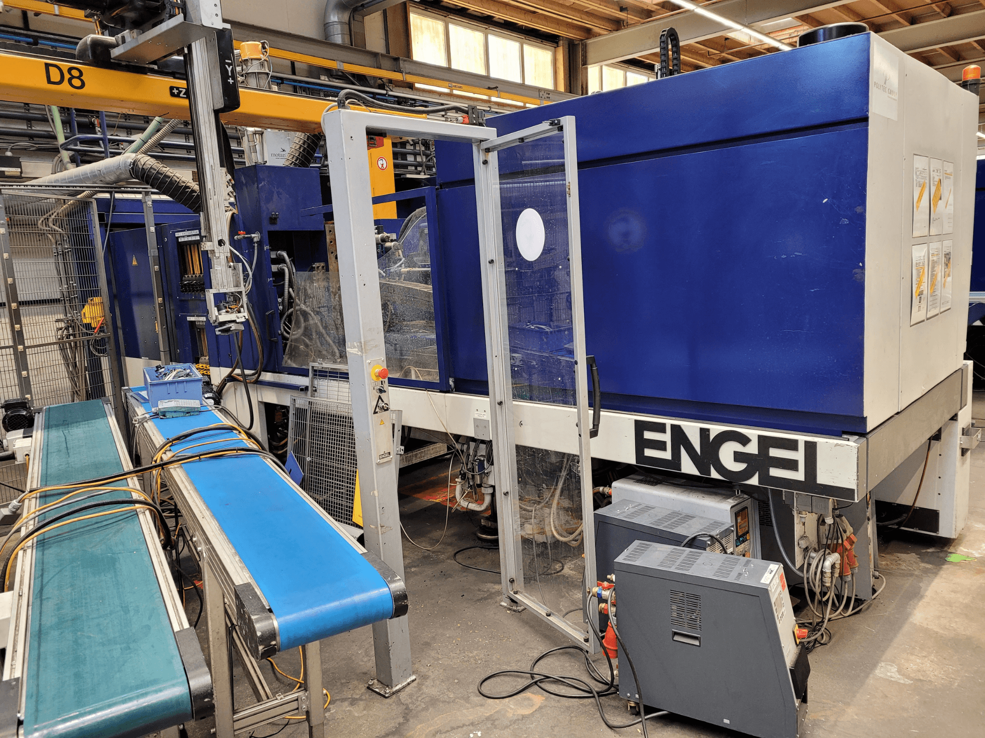 Máquina ENGEL azul com porta de segurança transparente, vista lateral, com painel de controlo, tapete transportador e ligações eléctricas visíveis.