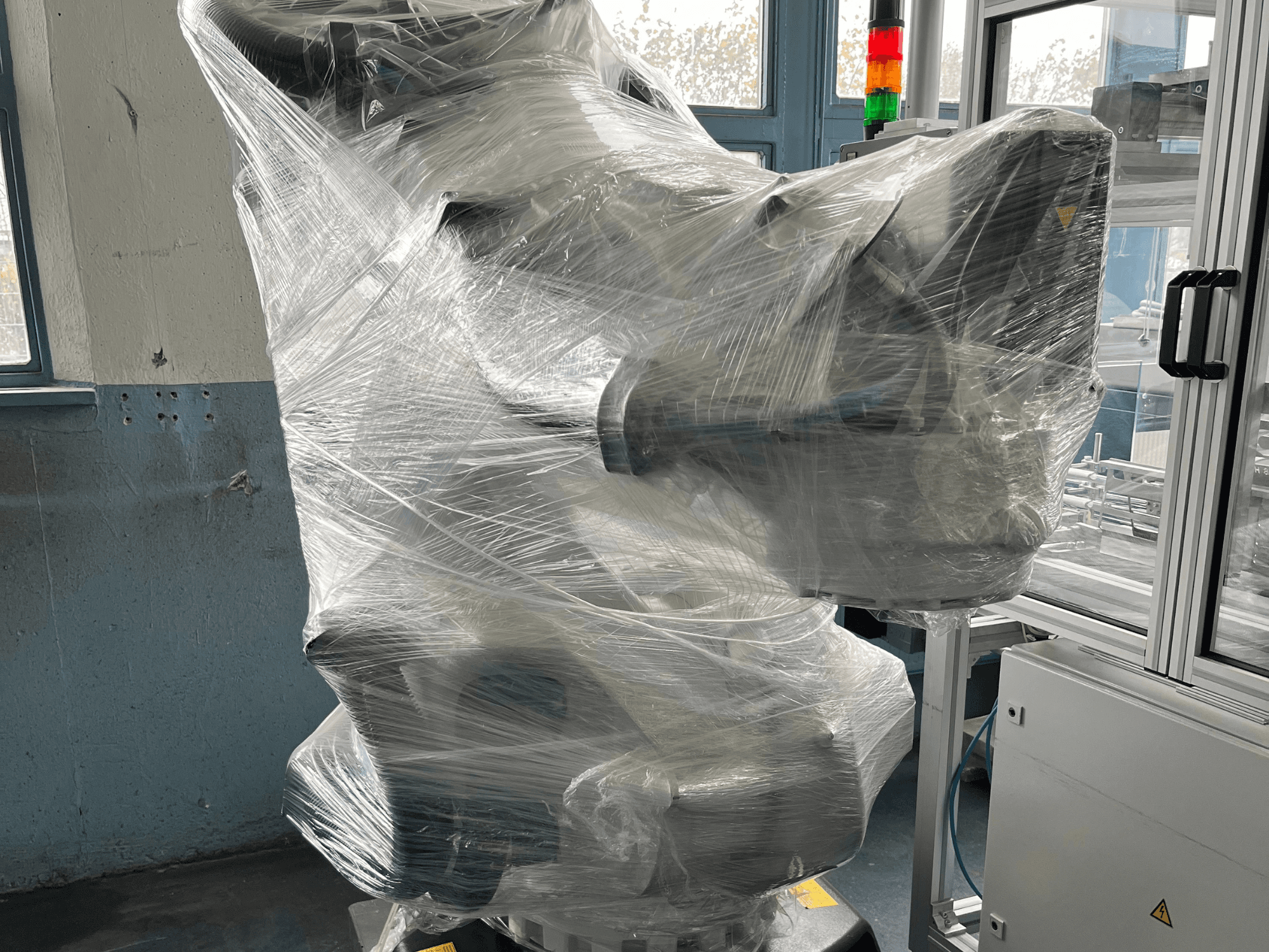 Braço robótico industrial envolto em plástico, vista frontal. Inclui unidade de controlo e luz indicadora. Marca não visível.