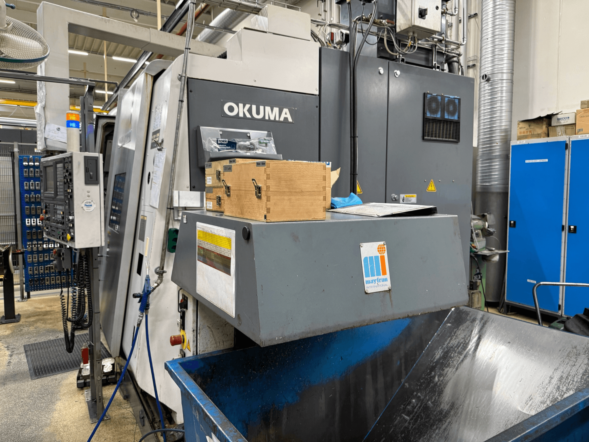 Máquina CNC Okuma na fábrica, vista frontal; apresenta painel de controlo, caixas de madeira e caixote do lixo azul com o logótipo Mayfran.