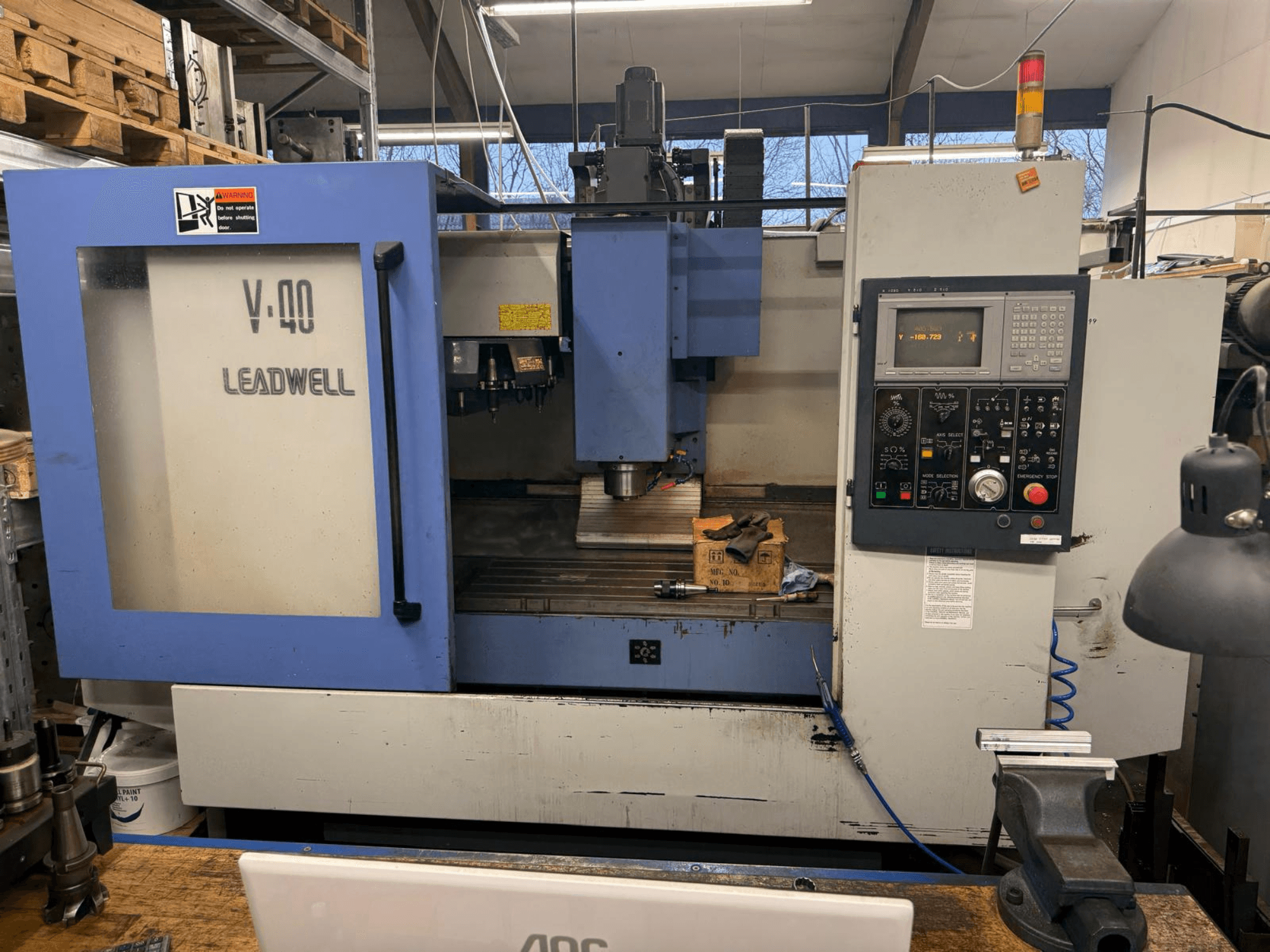 Fresadora LEADWELL V-40 CNC, vista frontal, com um painel de controlo, ferramentas de corte e um torno numa bancada de trabalho.