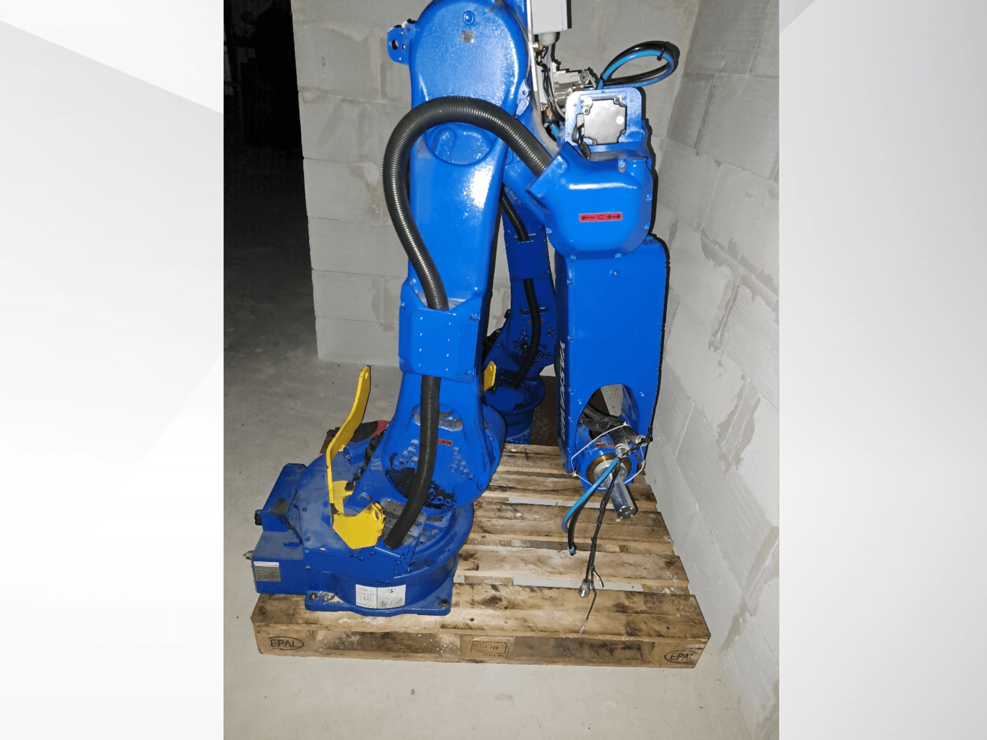 Braço robótico Yaskawa Motoman GP25, na cor azul, visto de lado sobre uma palete de madeira, com cabos e articulações mecânicas.