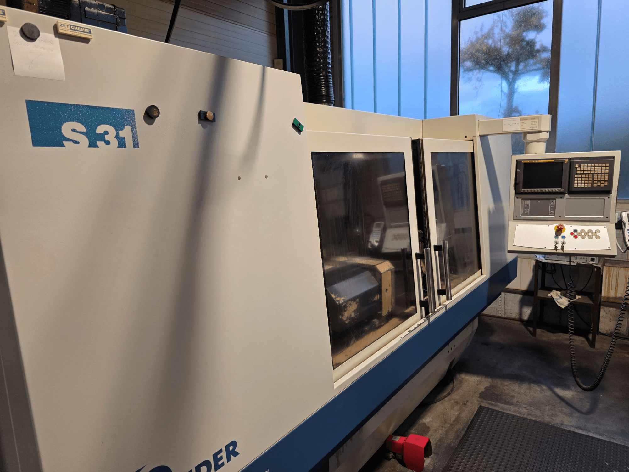Rectificadora CNC S31, vista frontal, painel de controlo com ecrã e botões, grande porta de vidro, ambiente industrial.