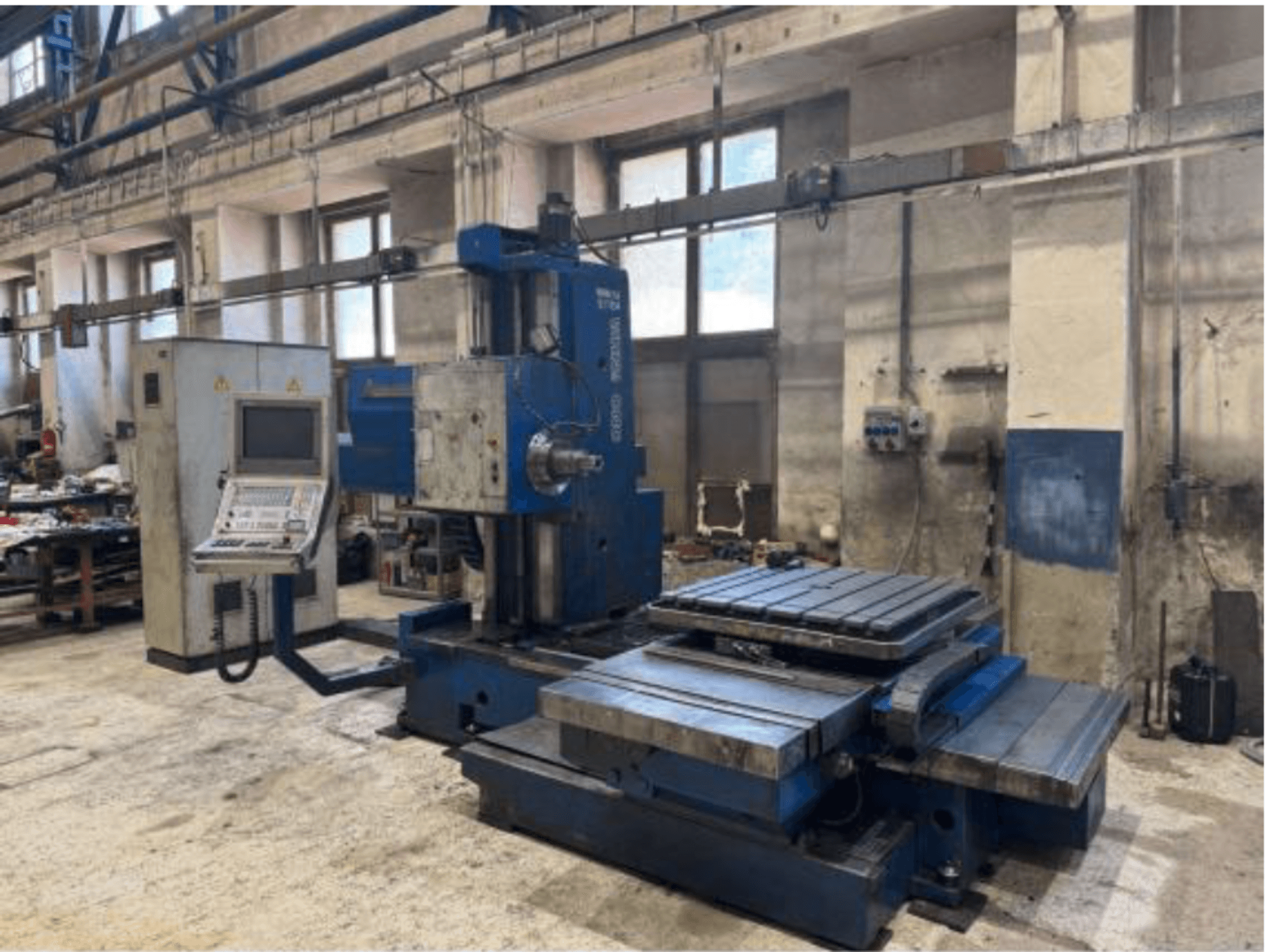 Vista frontal da máquina de mandrilagem CNC TOS WHN 9 B, com um painel de controlo e uma ampla mesa de trabalho, instalada num ambiente de oficina.