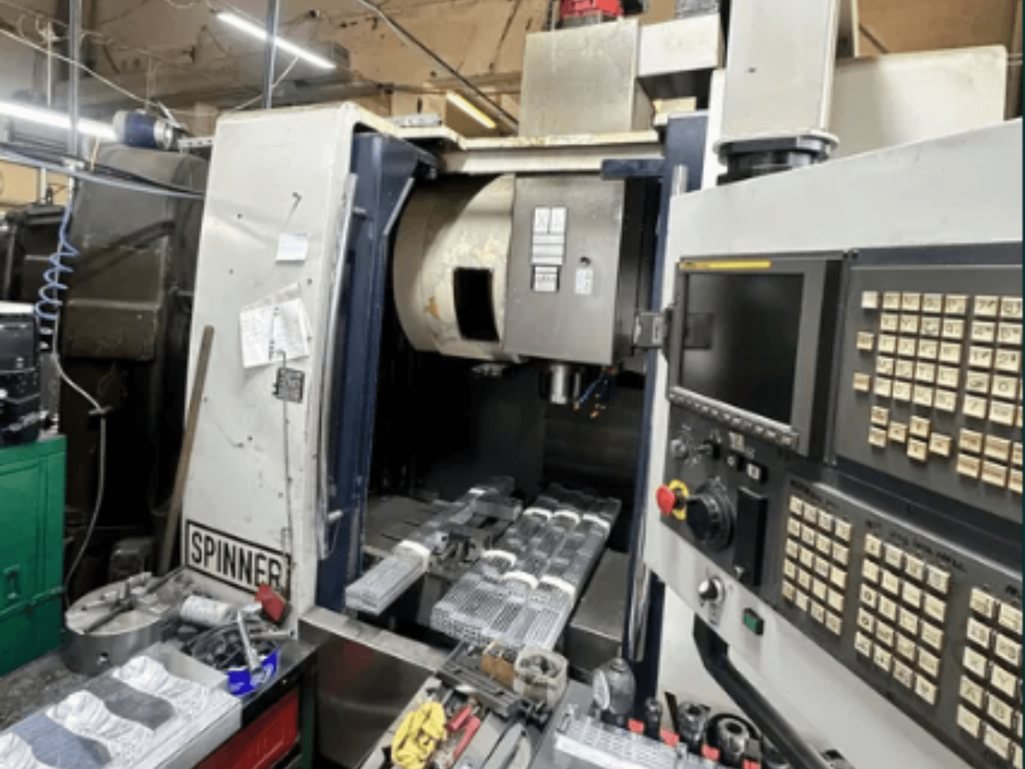 Máquina CNC da Spinner, vista frontal; apresenta uma interface de ecrã tátil, ferramentas na área de trabalho e peças maquinadas prontas para processamento.