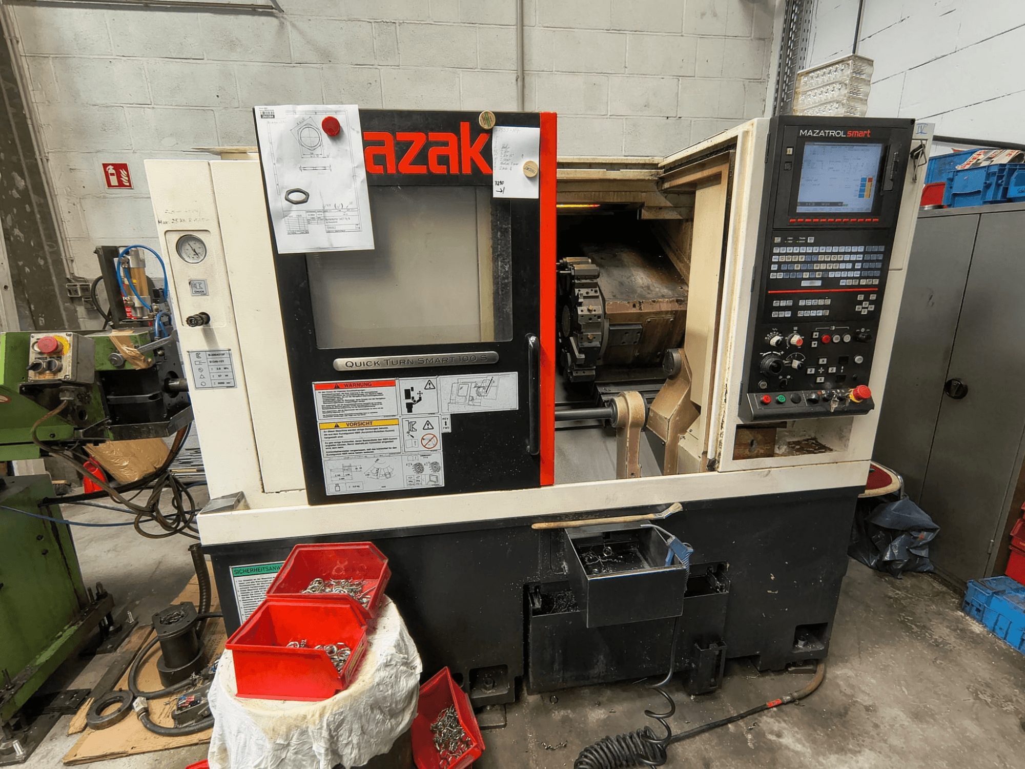Mazak Quick Turn Smart 100 S, vista lateral que mostra o painel de controlo, o tabuleiro de líquido de arrefecimento e os compartimentos de arrumação com parafusos.
