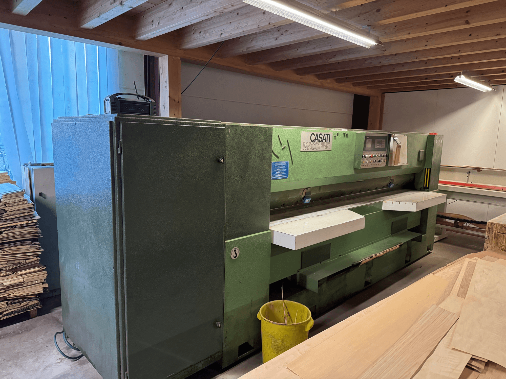 Máquina para trabalhar madeira CASATI verde numa oficina, vista de frente, com painel de controlo e superfícies de trabalho brancas, rodeada por pilhas de madeira.