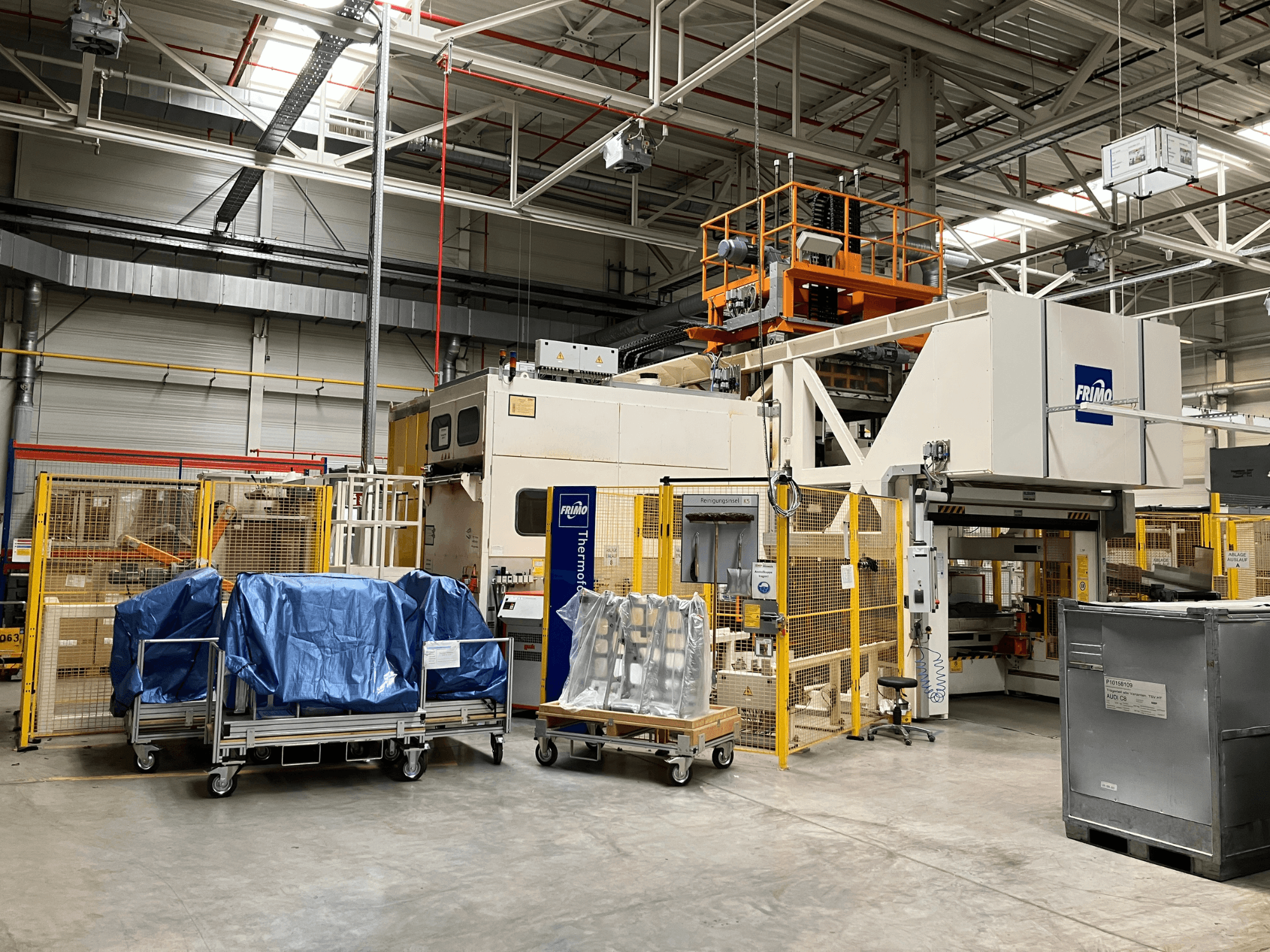 Máquina de processamento de plástico FRIMO FTL 80/200 Fullflex numa fábrica, vista frontal com lonas azuis e barreiras de segurança visíveis.