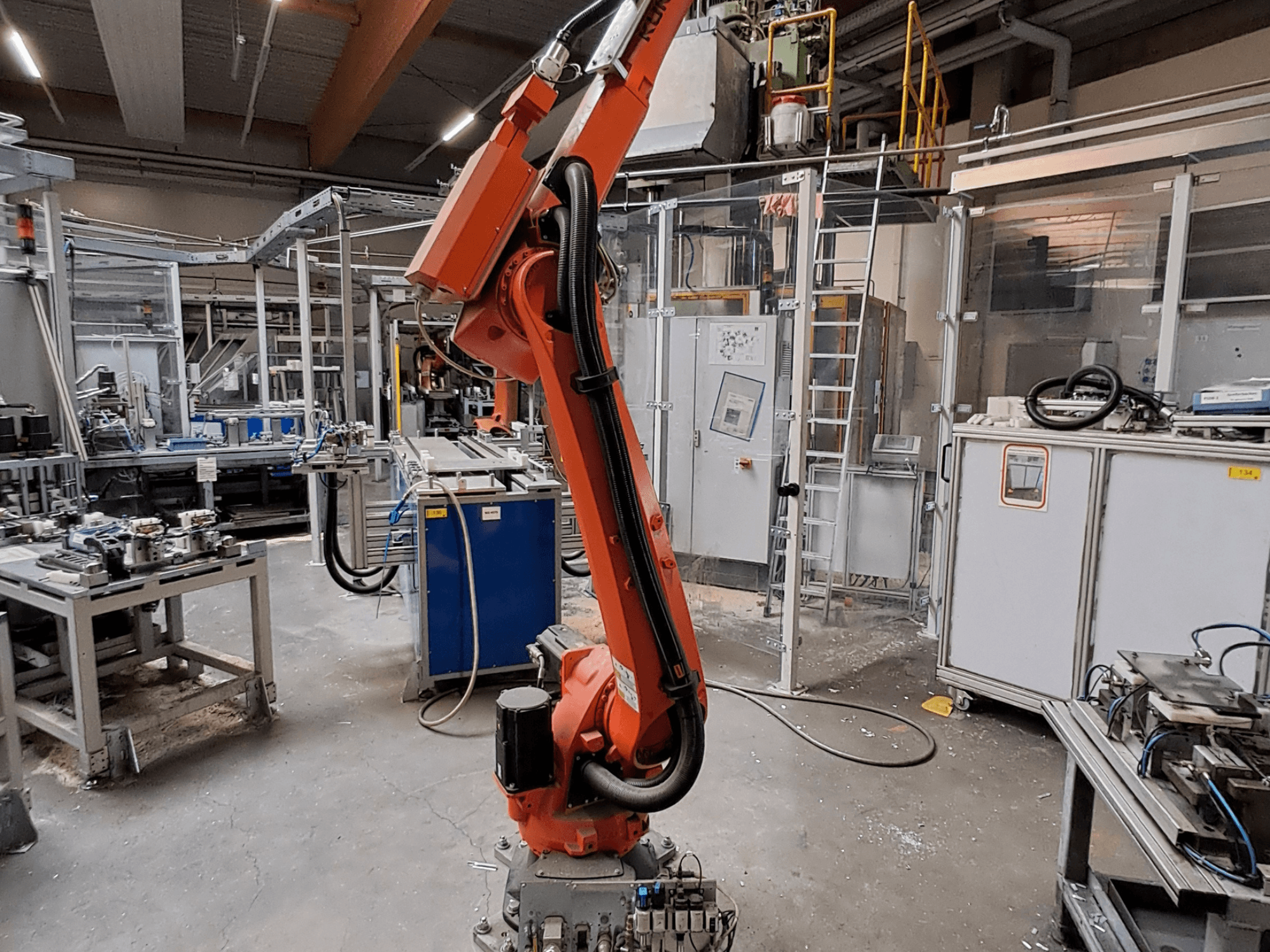 Braço robótico KUKA KR 26 R3100/SEL num ambiente industrial, visto de perfil, com equipamento operacional ao fundo.