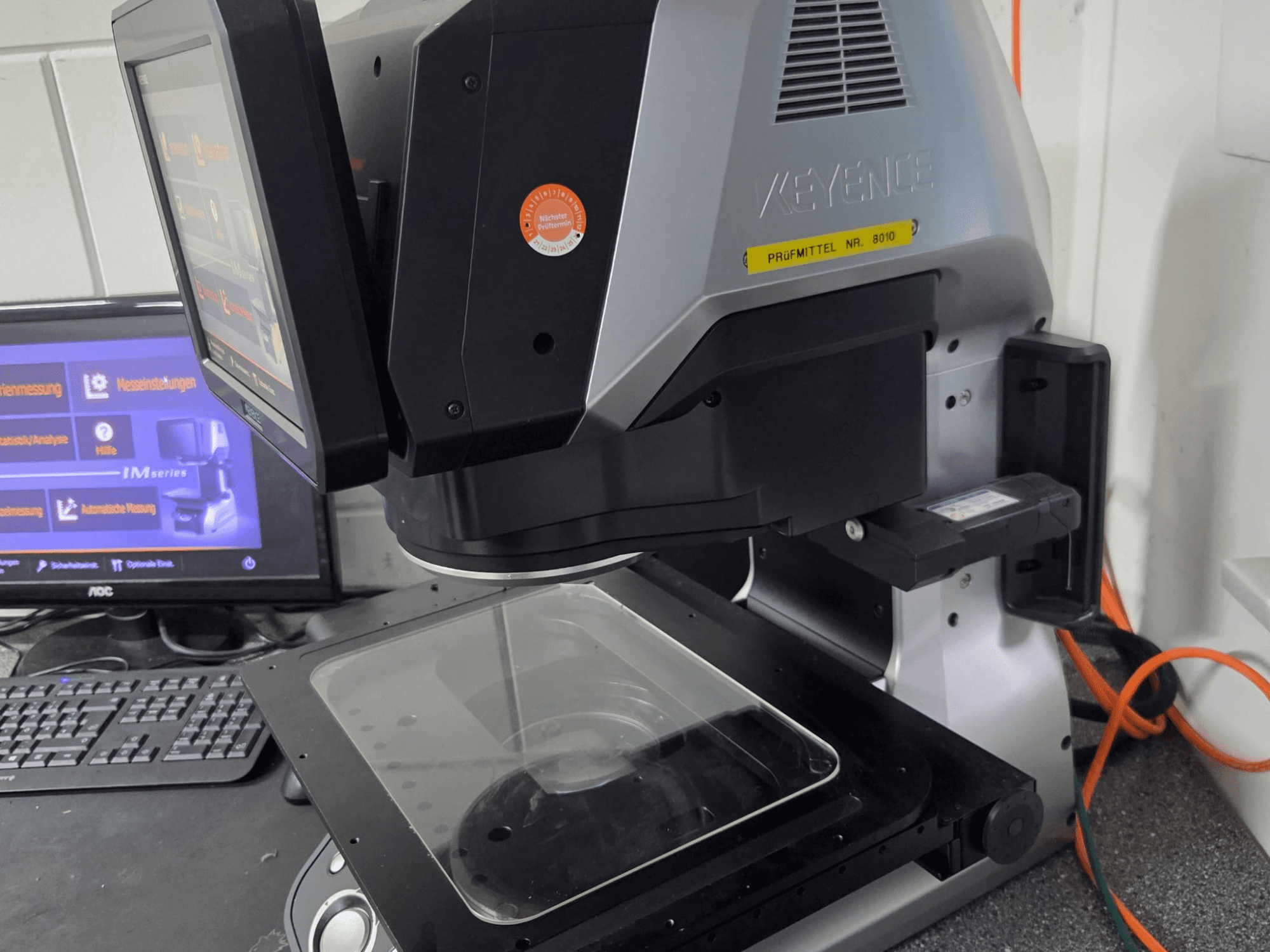 Máquina de medição ótica KEYENCE IM 7030, vista frontal com ecrã tátil e placa de medição em vidro.