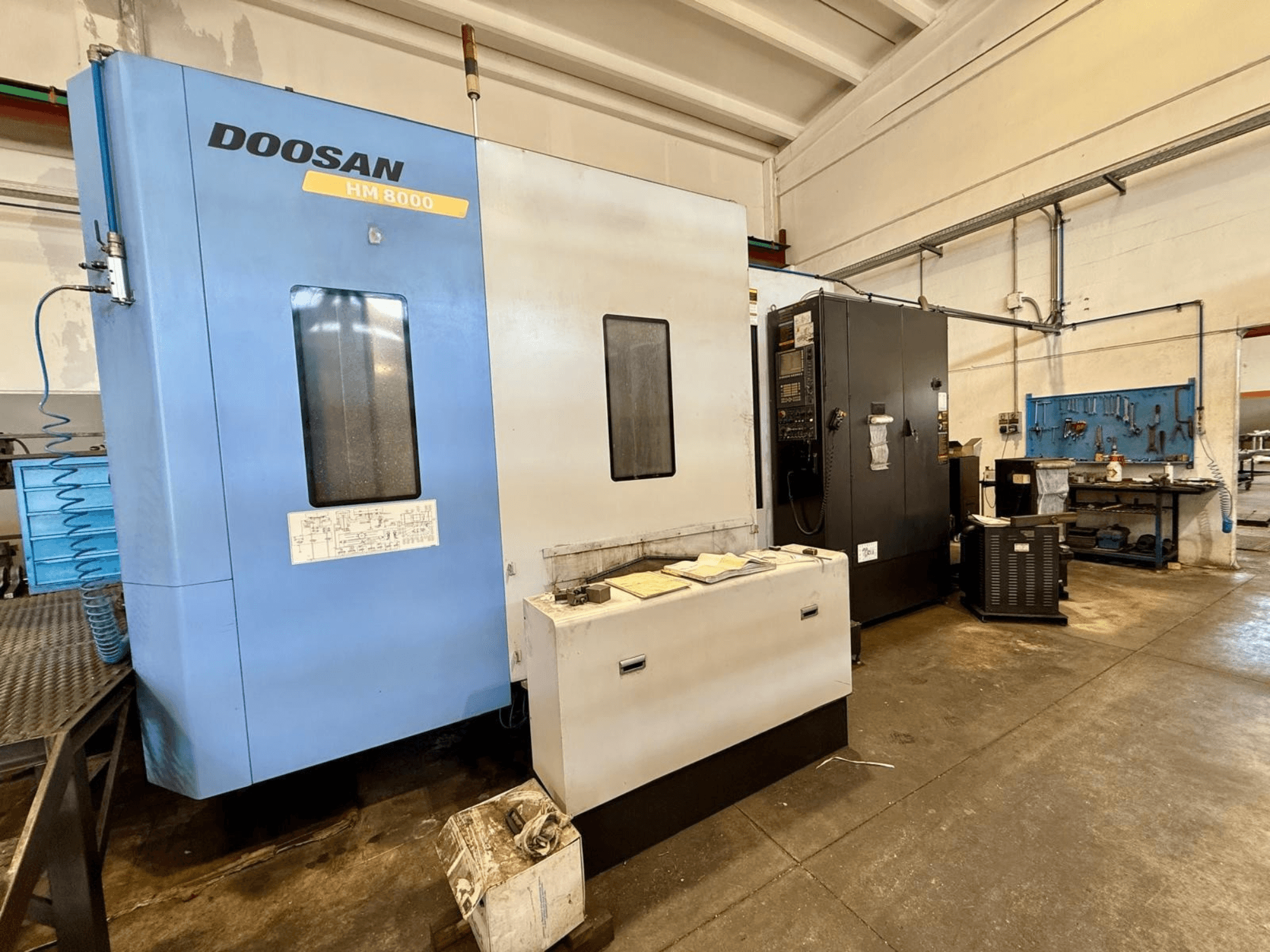 Centro de maquinagem horizontal DOOSAN HM 8000 em azul, vista frontal, mostrando o painel de controlo e o equipamento da oficina nas proximidades.