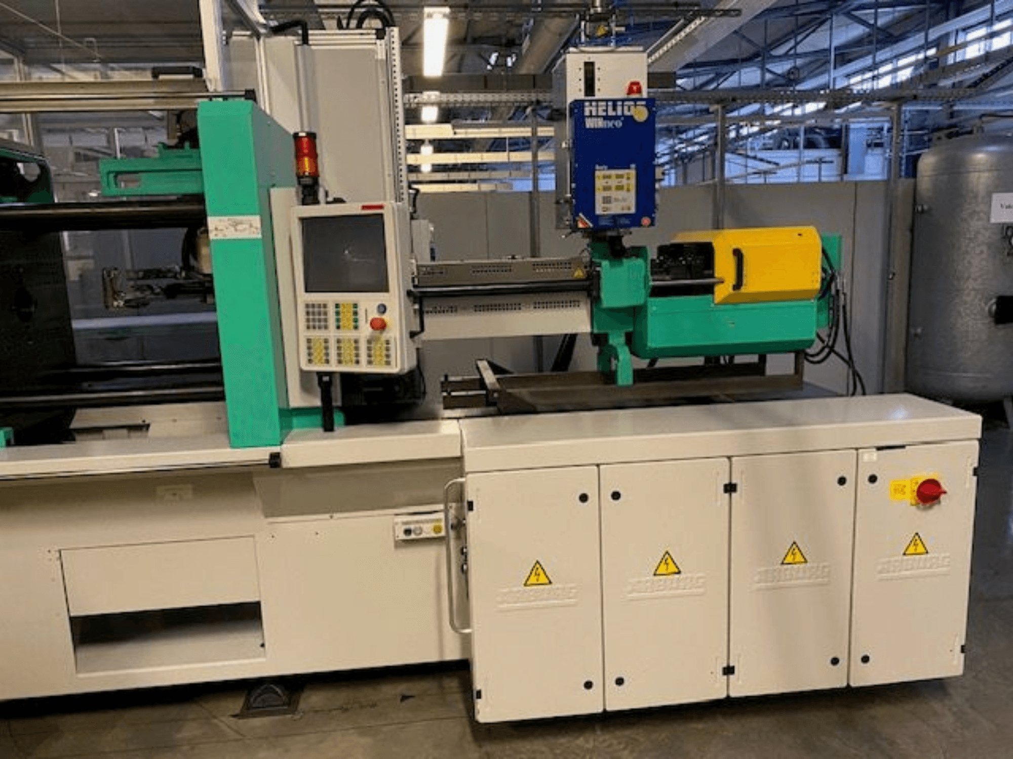 Máquina de moldagem por injeção industrial, modelo HELIO, vista frontal, com painel de controlo, protecções de segurança e botão de paragem de emergência.