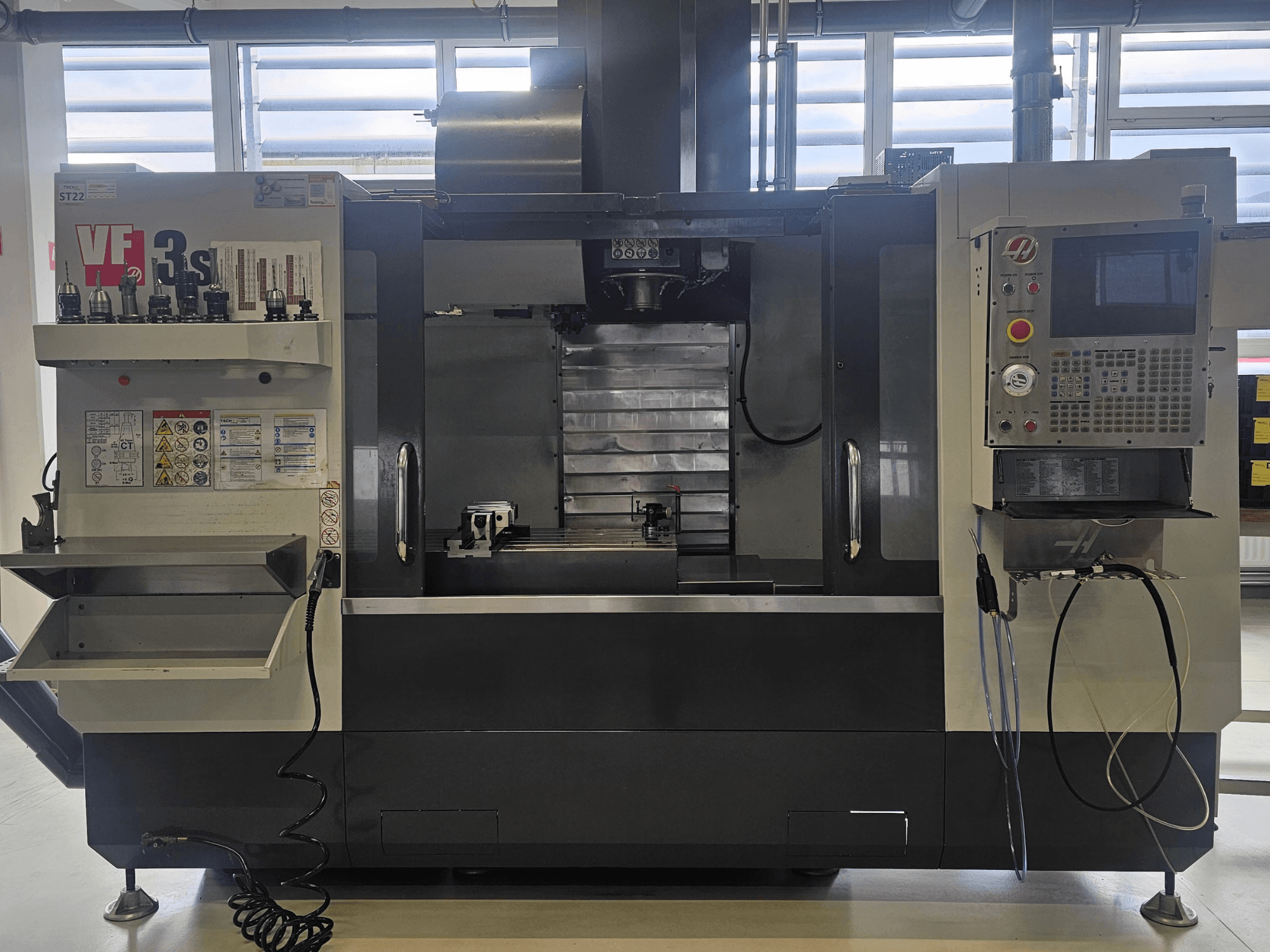 Fresadora CNC Haas VF3SS, vista frontal, com suportes de ferramentas, painel de controlo e tabuleiro de refrigeração vazio, com etiquetas de segurança visíveis.