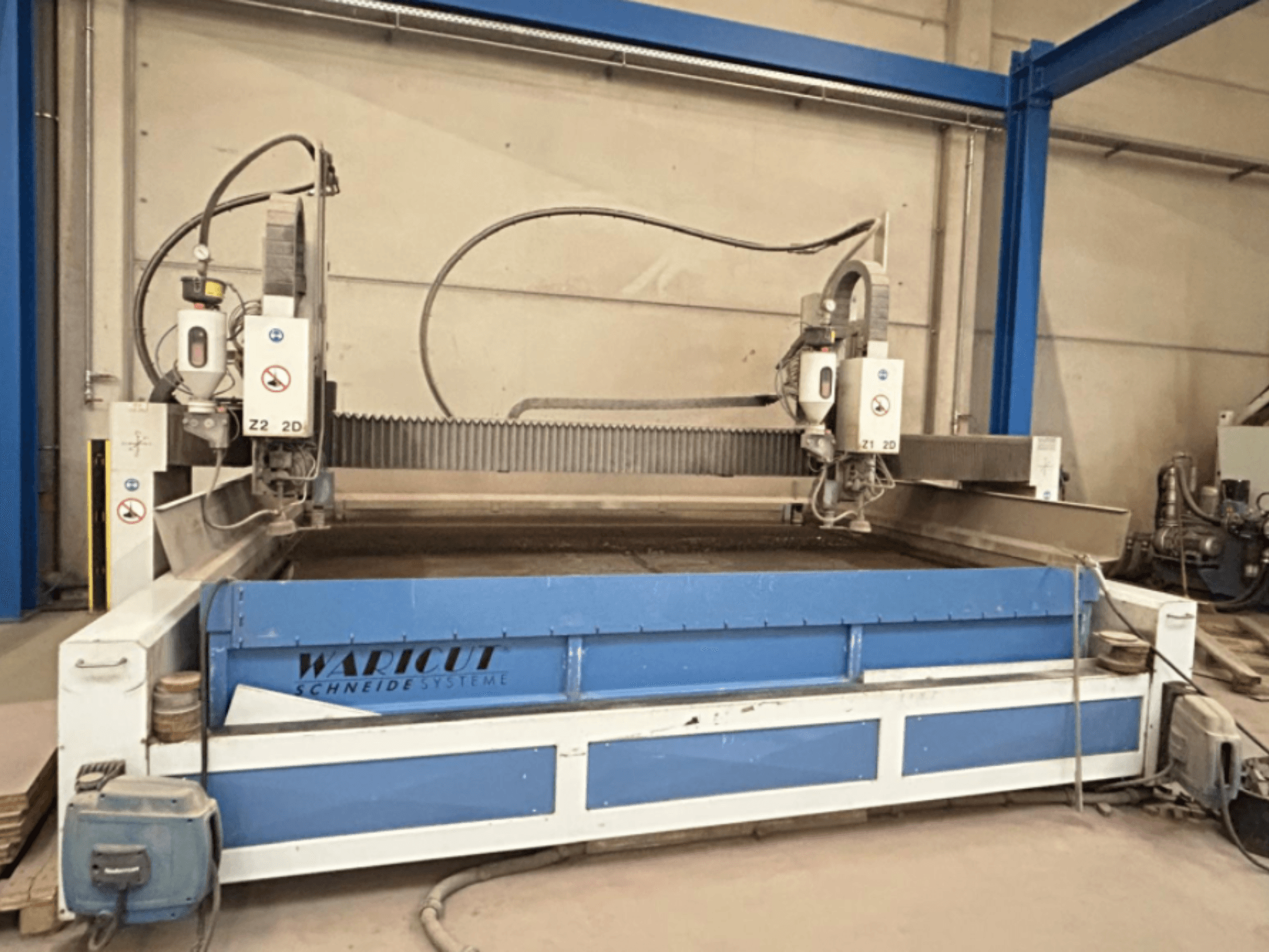 Máquina de corte por jato de água da Waricut, vista frontal; possui duas cabeças de corte e uma superfície de trabalho azul numa oficina.