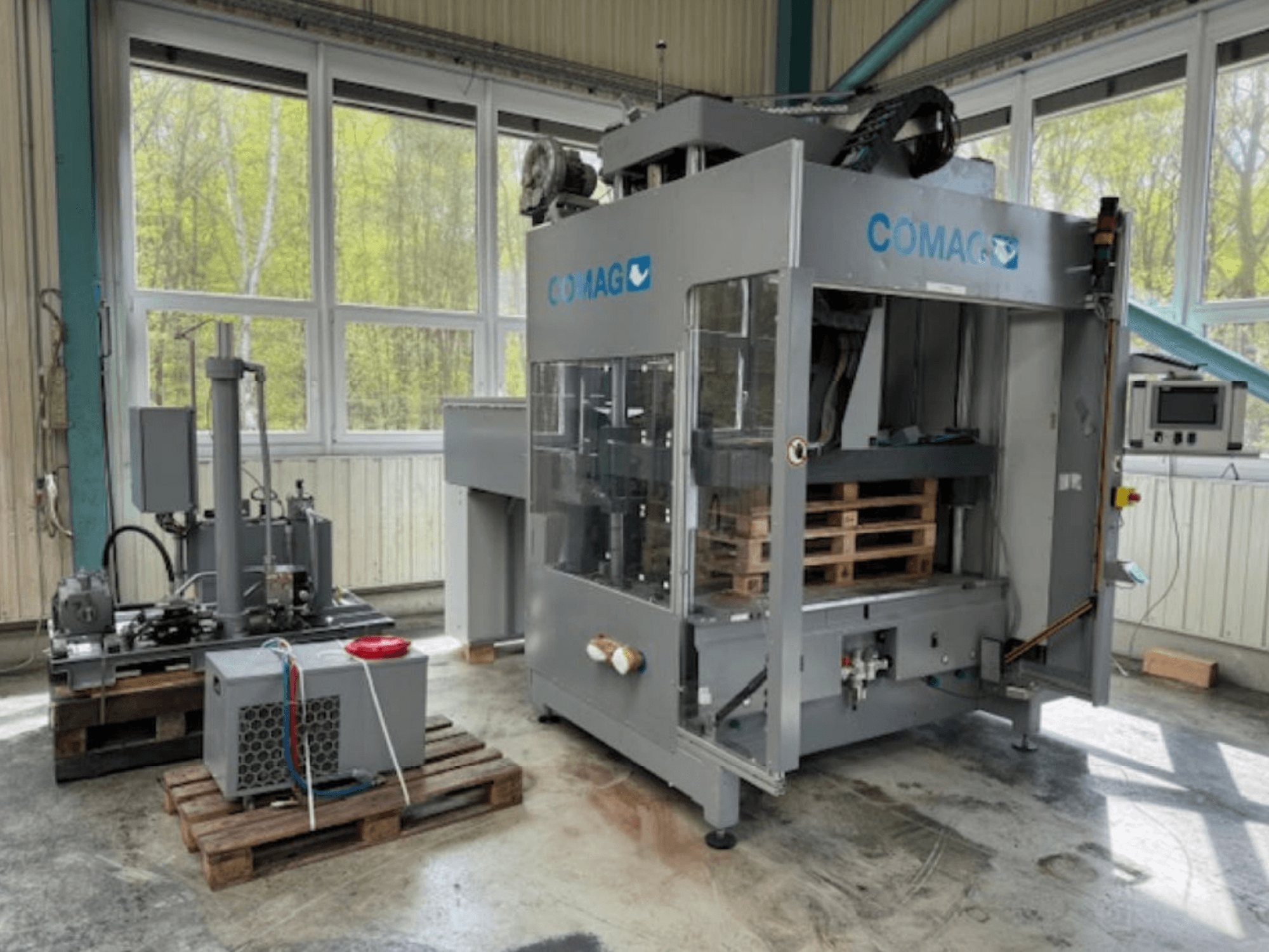 Máquina de embalar paletes COMAG em ambiente industrial, vista frontal; paletes e componentes hidráulicos visíveis.