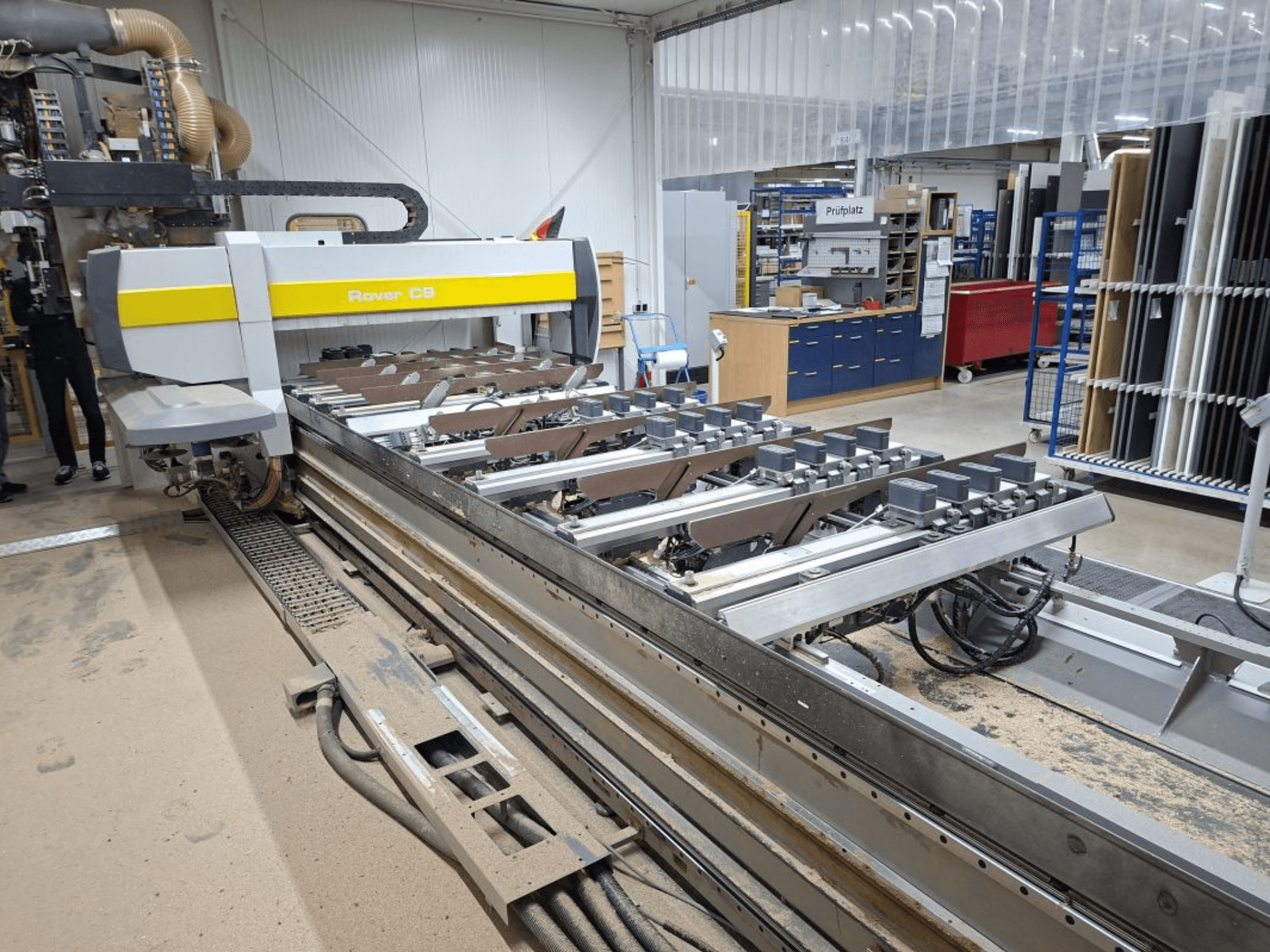 Máquina CNC Rover C9 numa oficina; a vista frontal mostra as ferramentas de corte e o sistema de transporte, com acumulação de pó no chão.