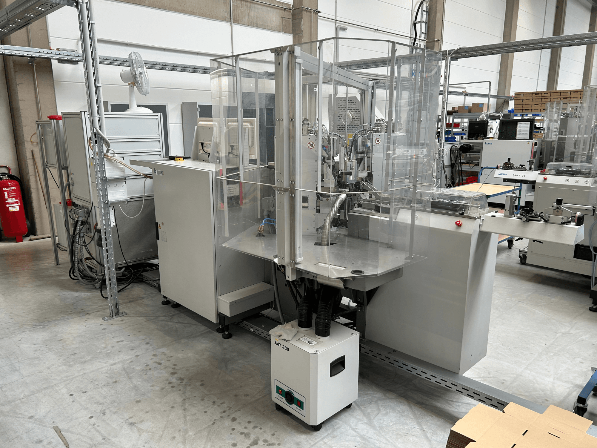 Equipamento de automação KOMAX Alpha 433 L num ambiente fabril. Apresenta uma cabina de segurança transparente e vários painéis de controlo.