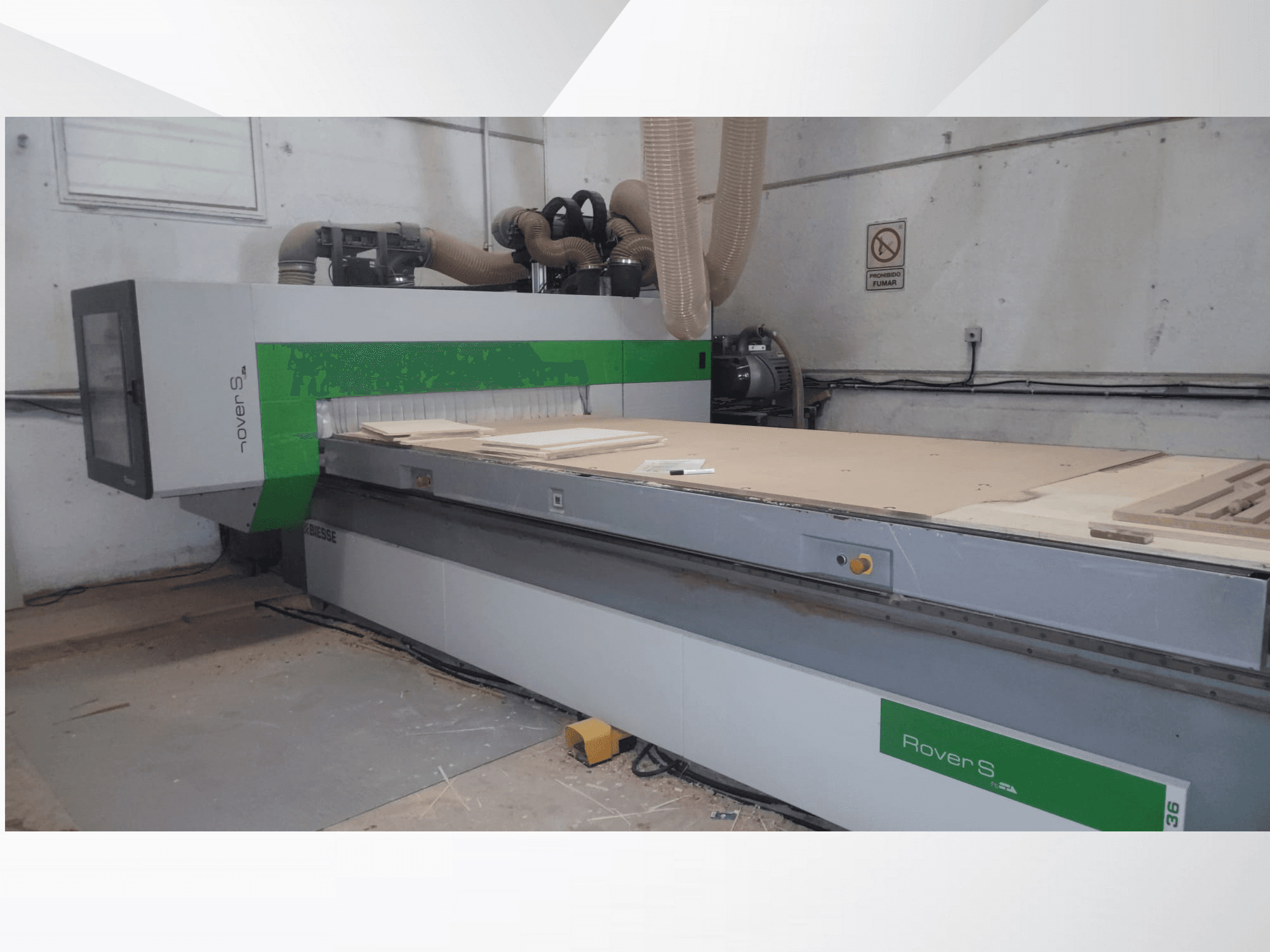 Vista frontal da máquina CNC Biesse Rover S, com uma grande superfície de trabalho com painéis de madeira e mangueiras de extração de poeiras.