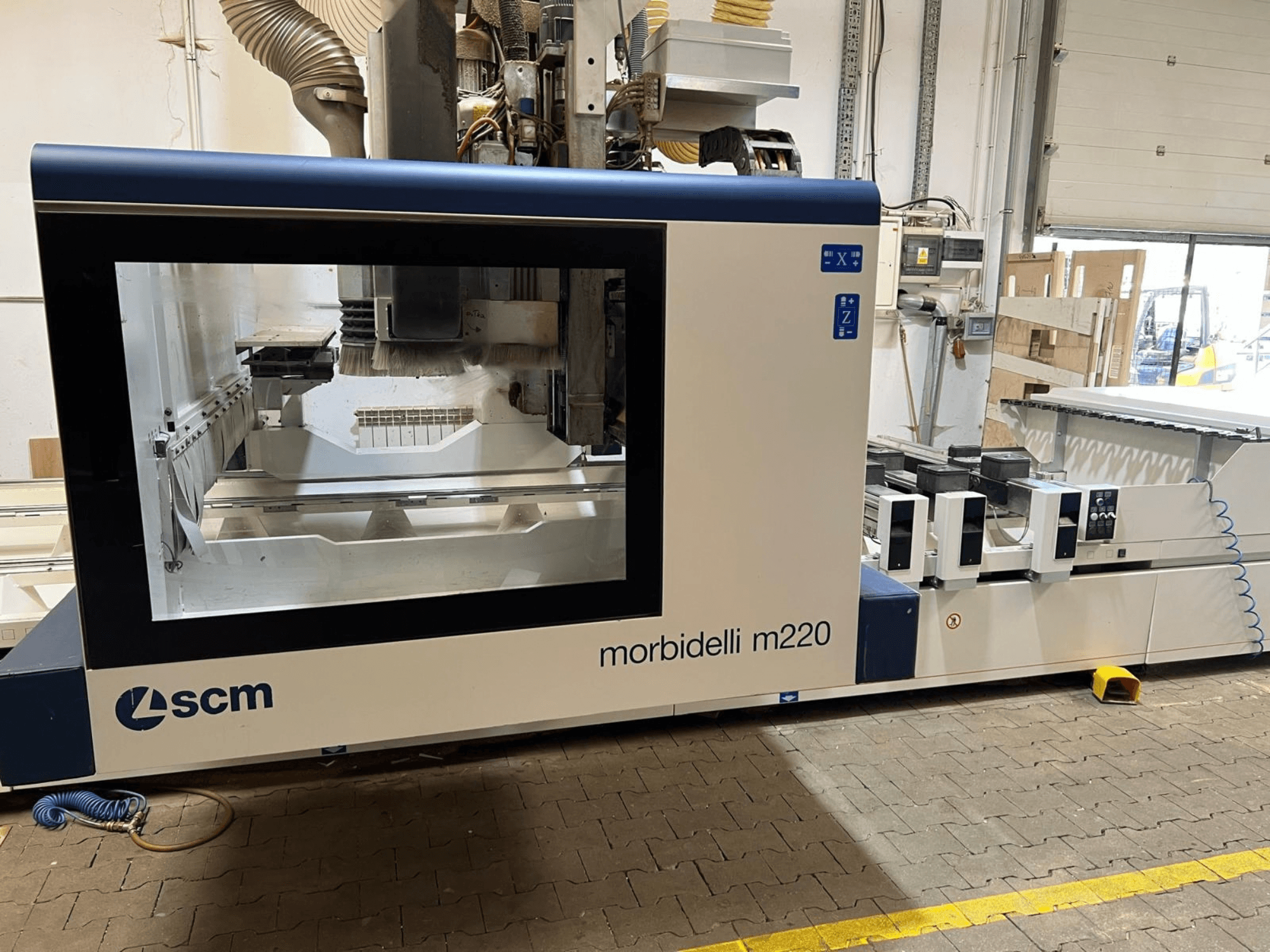 Vista frontal do Centro de Processamento CNC SCM Morbidelli M220, que apresenta uma ampla janela, um painel de controlo e uma área de trabalho.