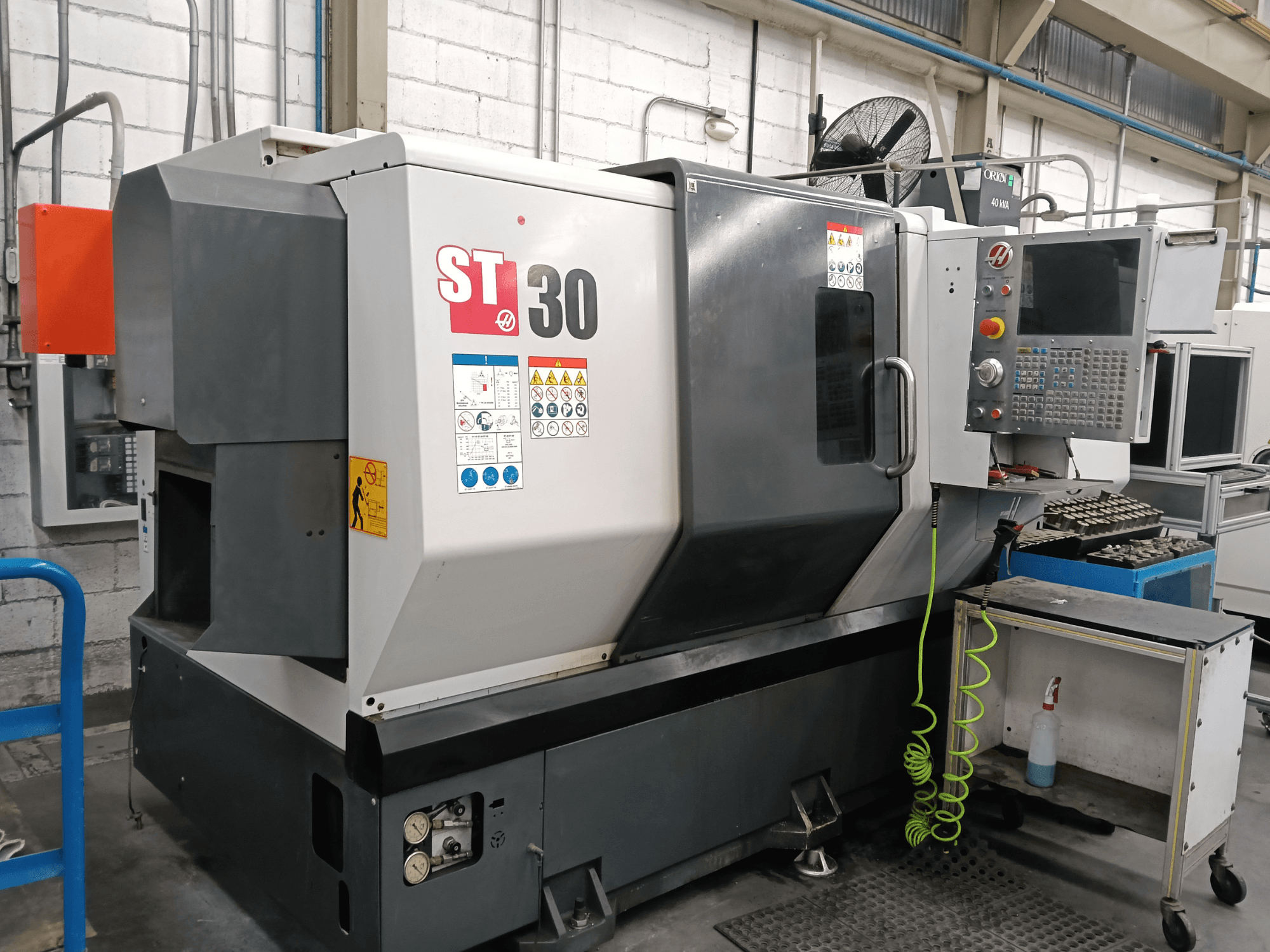 Texto alternativo: Vista frontal do torno horizontal HAAS ST-30 com painel de controlo, etiquetas de segurança e ferramentas num carrinho nas proximidades.