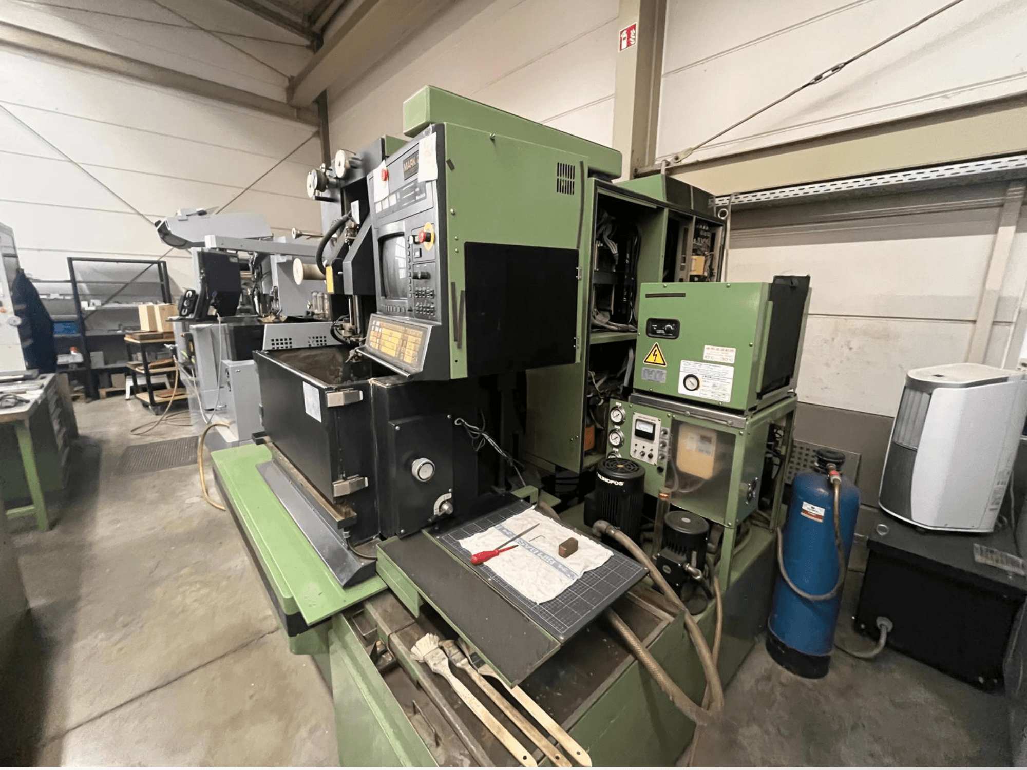 Máquina industrial verde com painel de controlo, vista lateral; inclui componentes hidráulicos e superfície de trabalho com ferramentas.