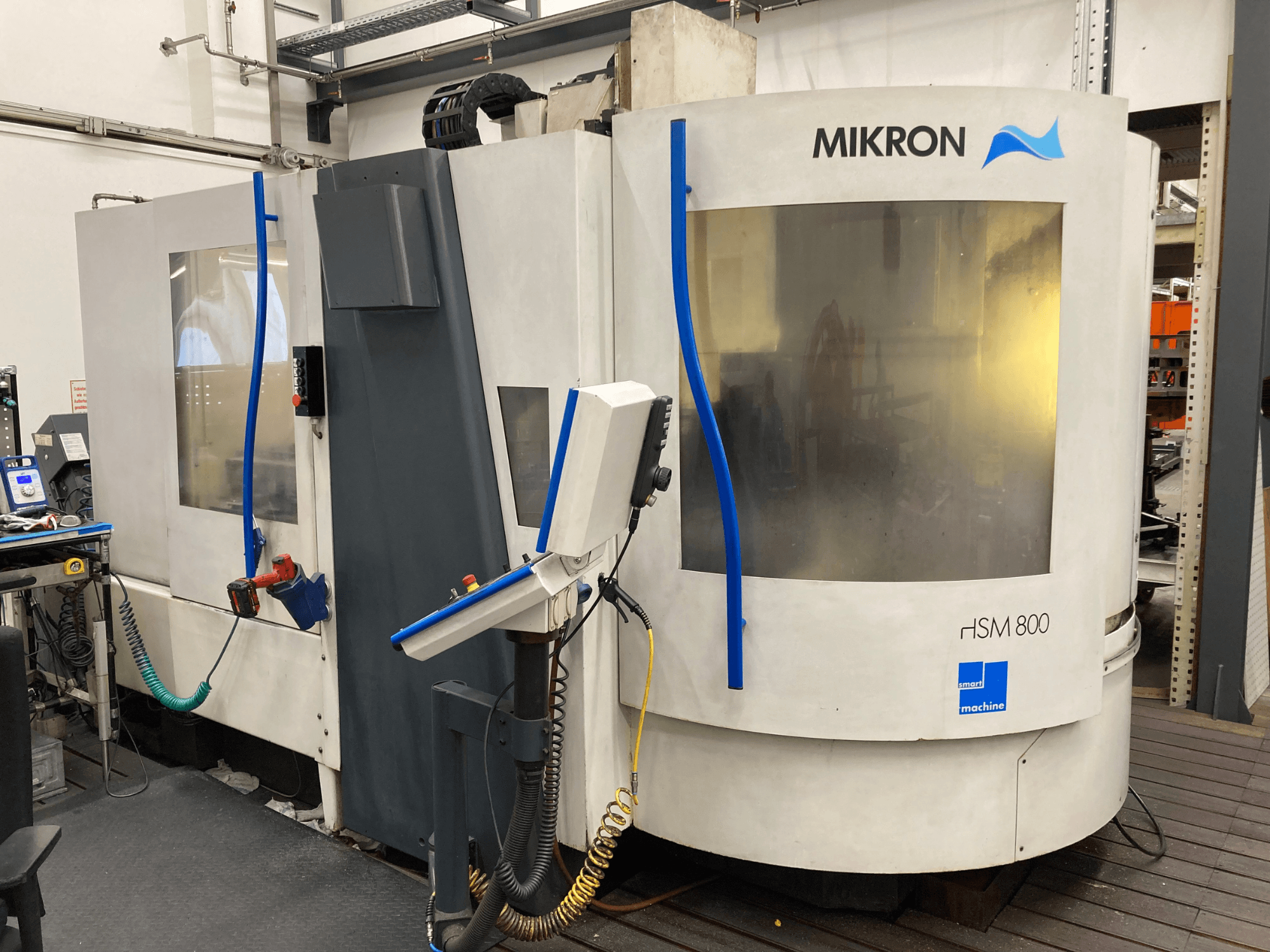Mikron HSM 800, vista lateral de um centro de maquinação CNC com painel de controlo e suporte de ferramentas, mangueiras e cabos visíveis.