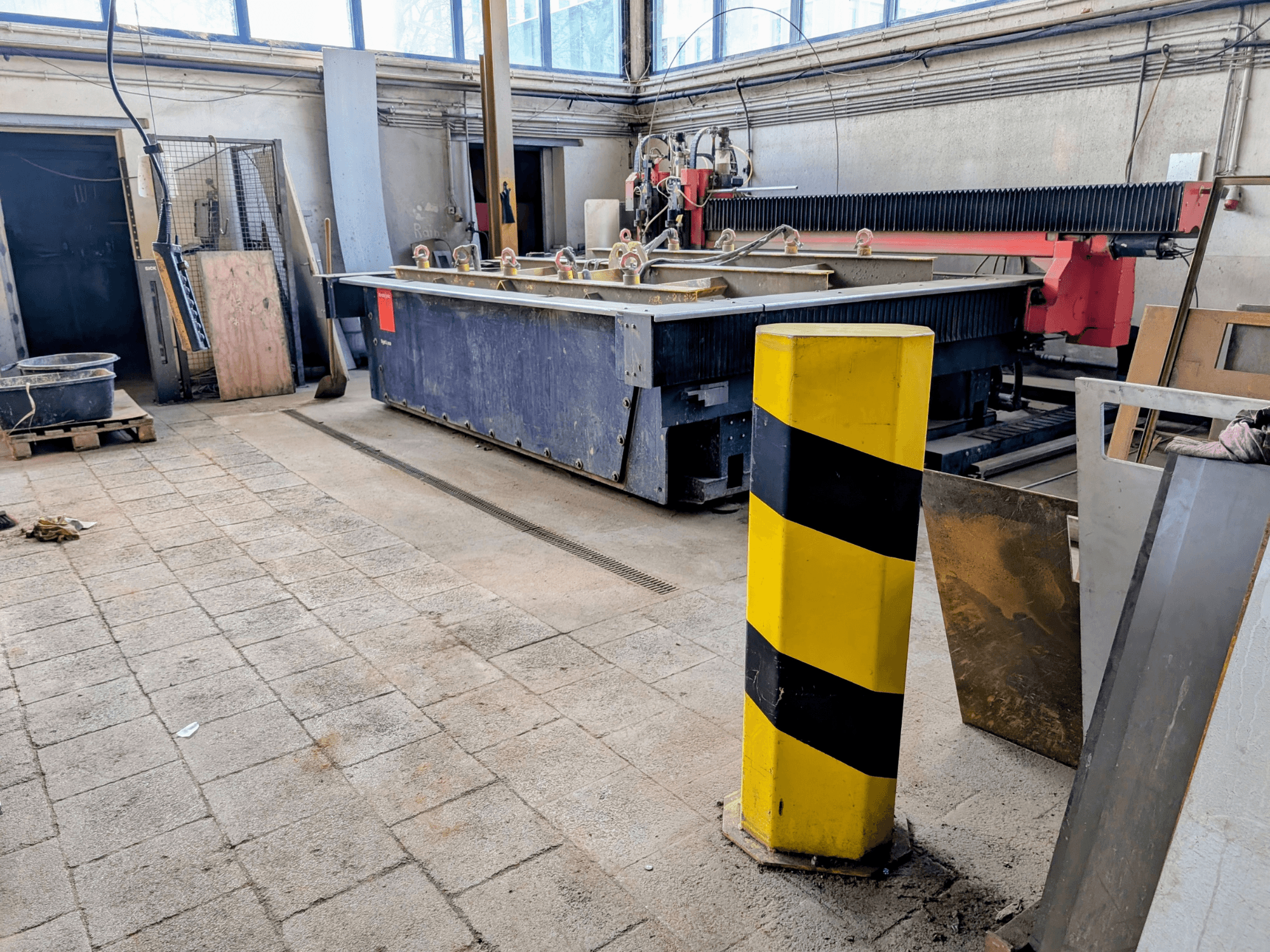 Máquina de corte por jato de água Bystronic Byjet L 2030 numa oficina, vista lateral, com um poste de segurança amarelo e materiais circundantes.