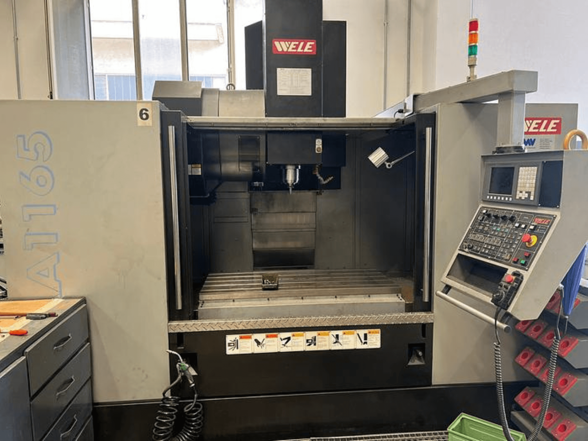 Centro de maquinação CNC WELE A165, vista frontal, com painel de controlo, suporte de ferramentas e mesa de trabalho visíveis. Ambiente industrial.