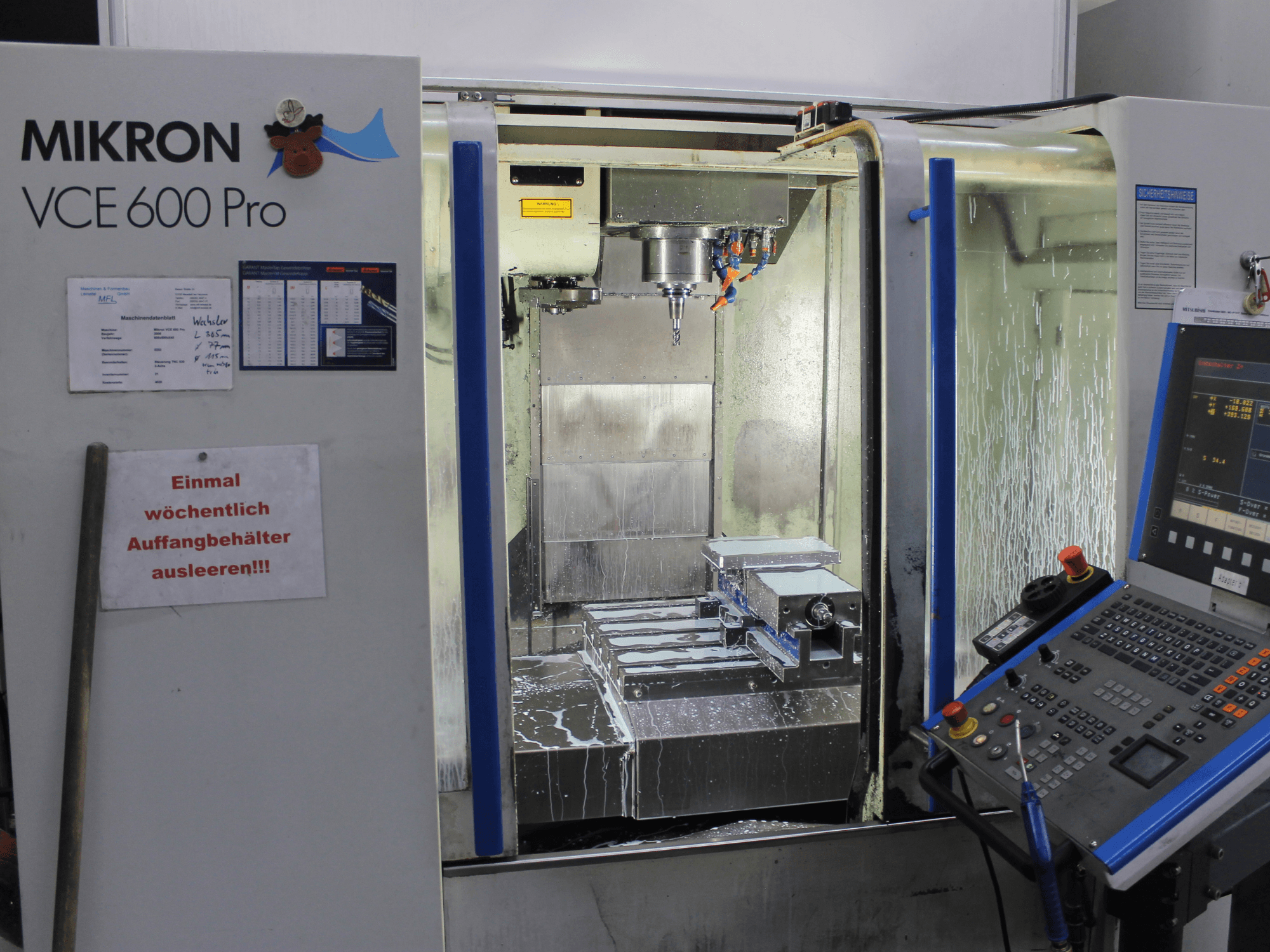 Centro de maquinação Mikron VCE 600 Pro, vista frontal, com ferramentas de corte, pulverização de líquido de refrigeração e painel de controlo com botões.