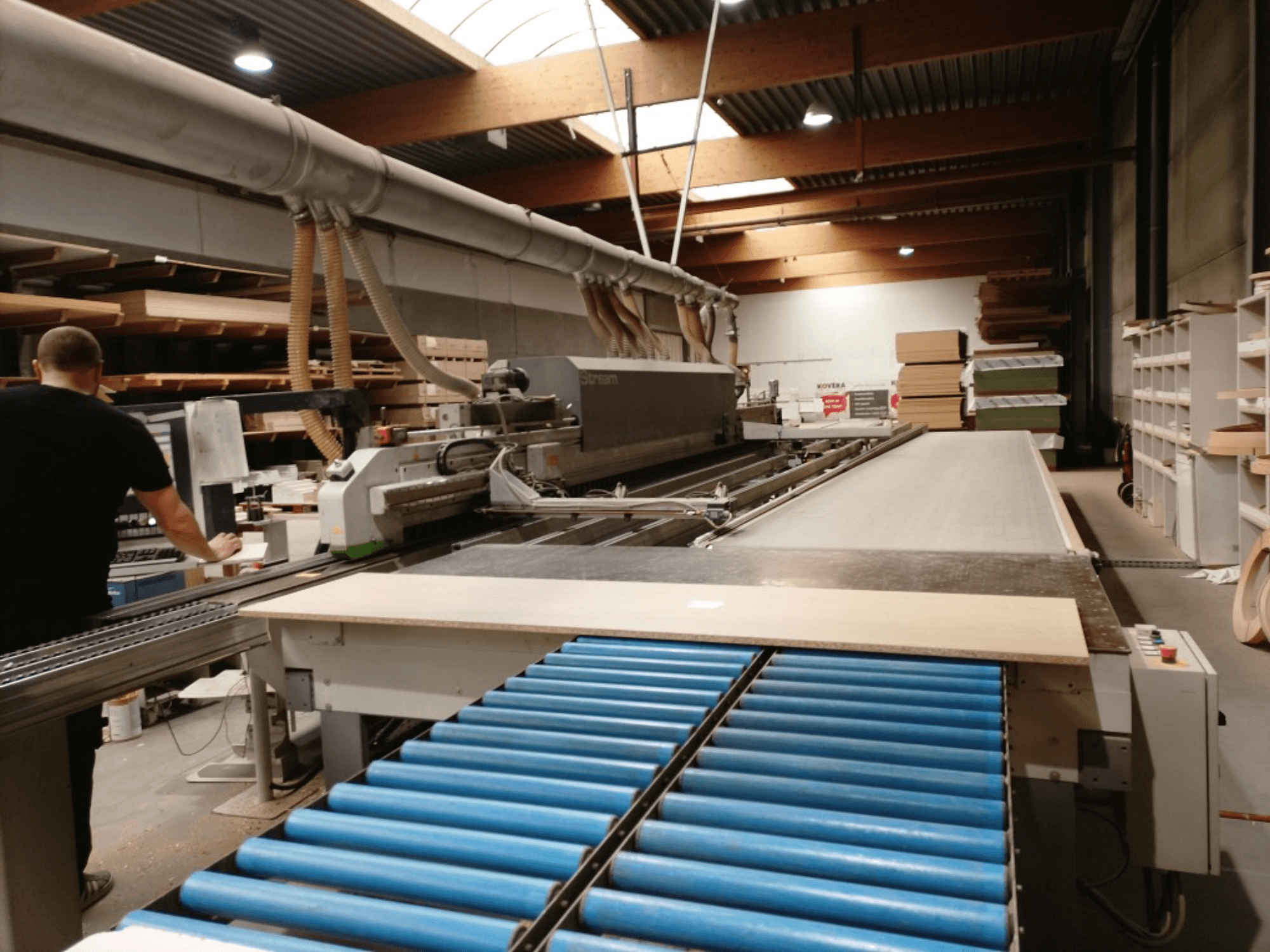 BIESSE Edge Stream B1, vista lateral mostrando o operador, os rolos azuis e o painel de madeira no transportador.