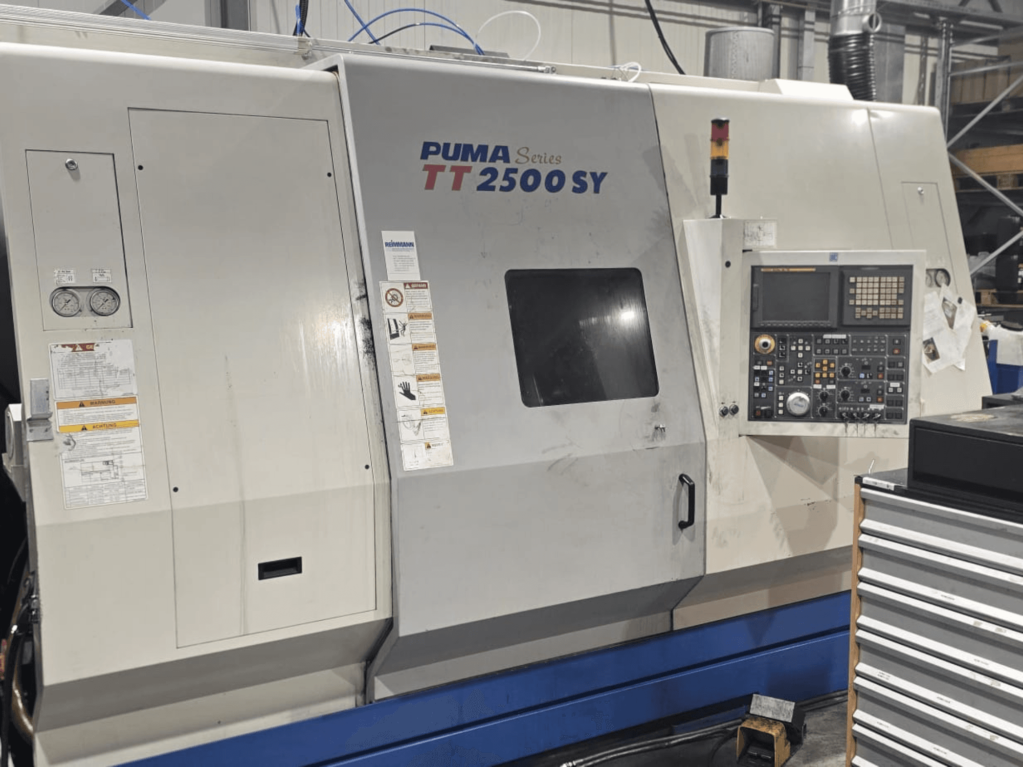 Torno CNC PUMA Série TT 2500 SY, vista frontal; painel de controlo com mostradores e botões, manómetros e etiquetas de operação visíveis.