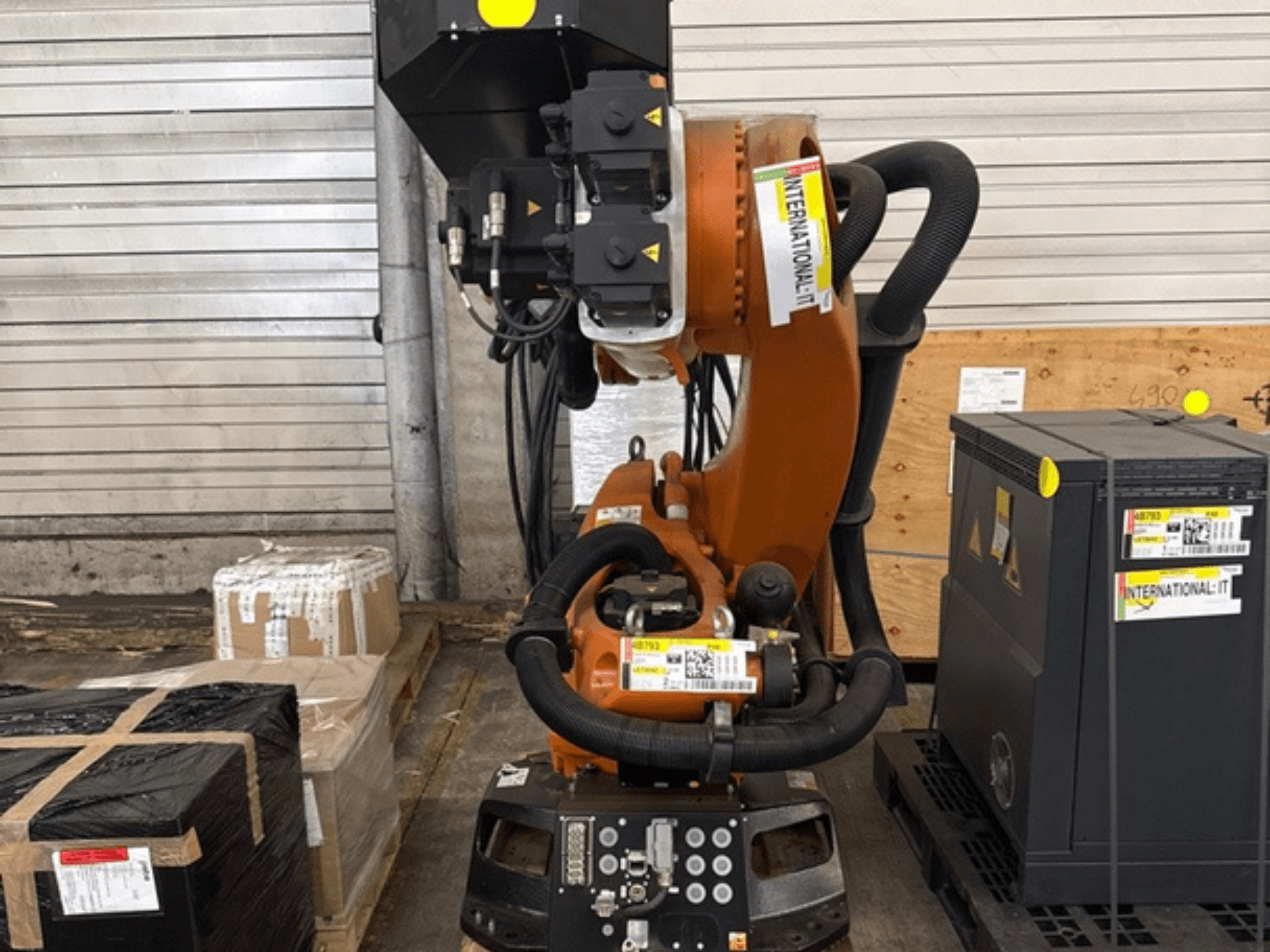 Braço robótico adicional KUKA KR 210 R2700, visto de frente, exposto numa palete ao lado de embalagens e equipamento.
