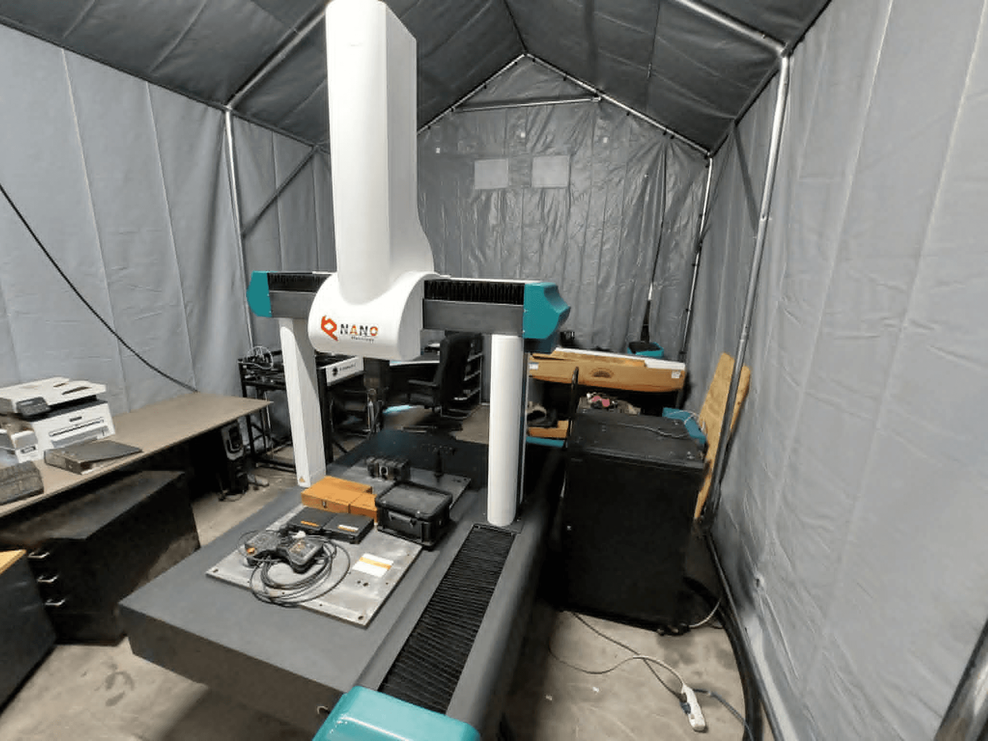 NANO METROLOGY Micr 7106 CMM numa tenda; vista frontal mostrando o braço da máquina e a plataforma de medição com acessórios.
