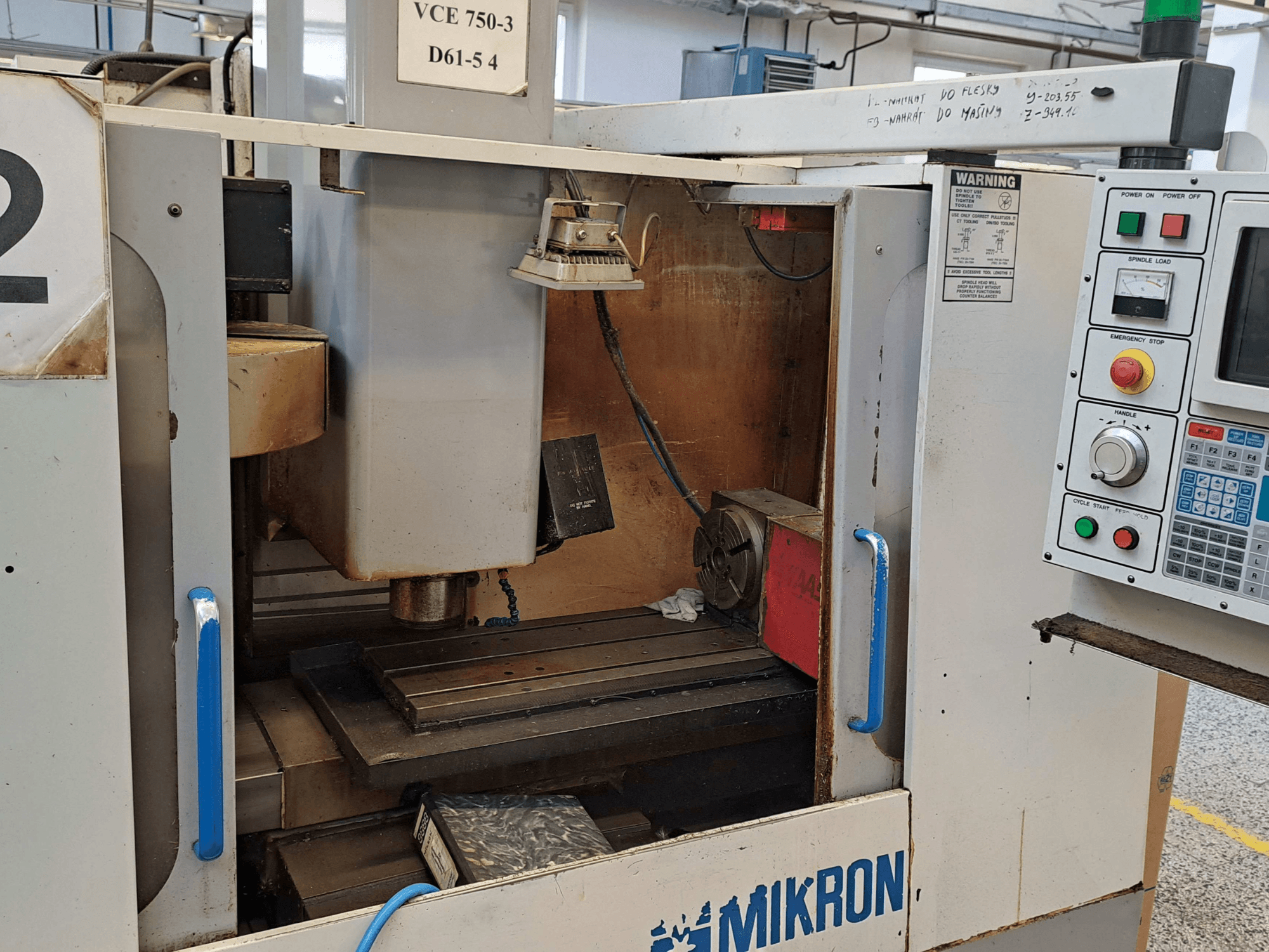 Vista frontal de uma fresadora Mikron VCE 750-3, mostrando o fuso, o painel de controlo e a mesa de trabalho com detalhes técnicos.