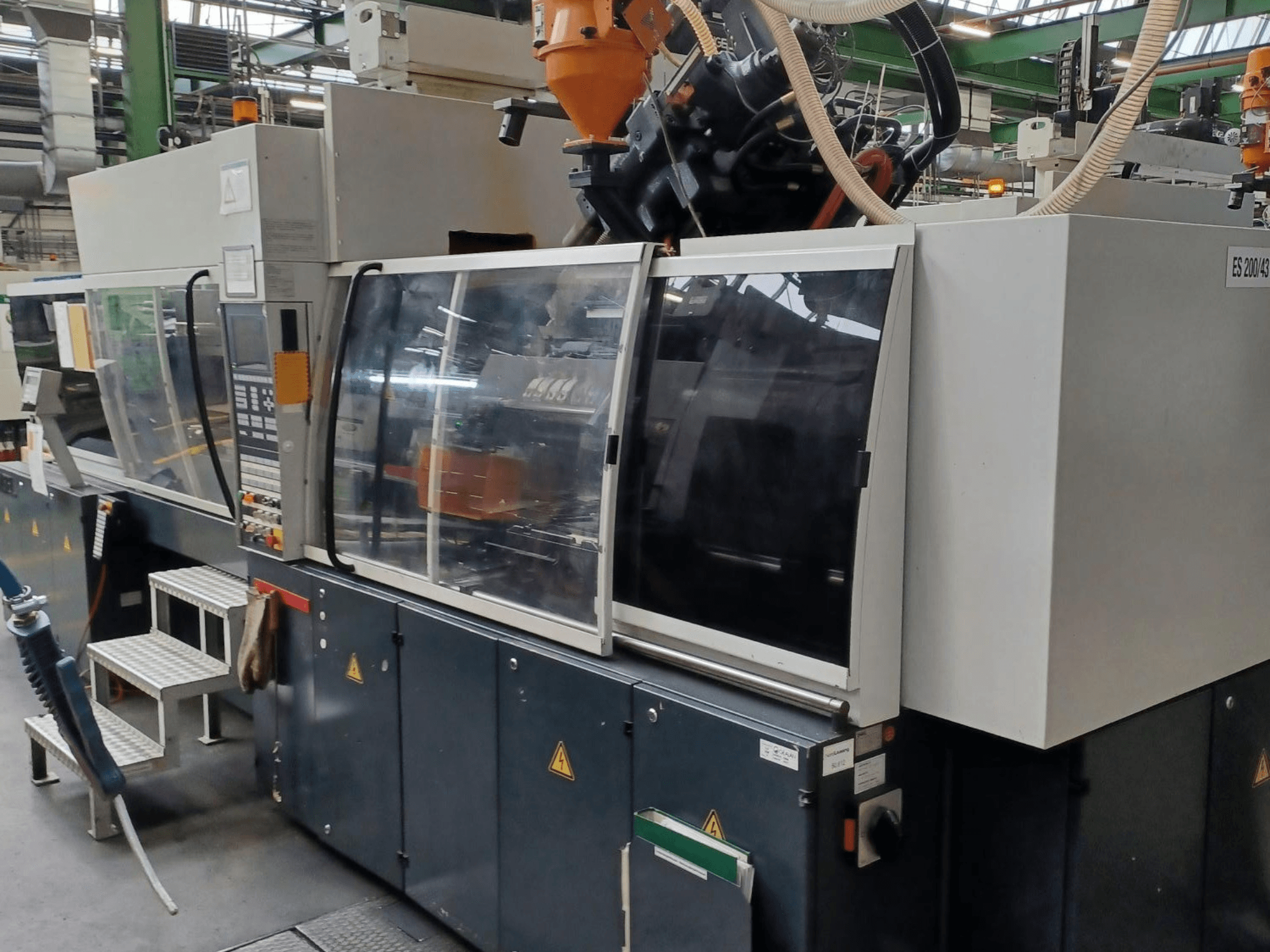 Vista frontal de uma máquina de moldagem por injeção de plástico, de marca e modelo desconhecidos, com portas de segurança transparentes e painel de controlo.