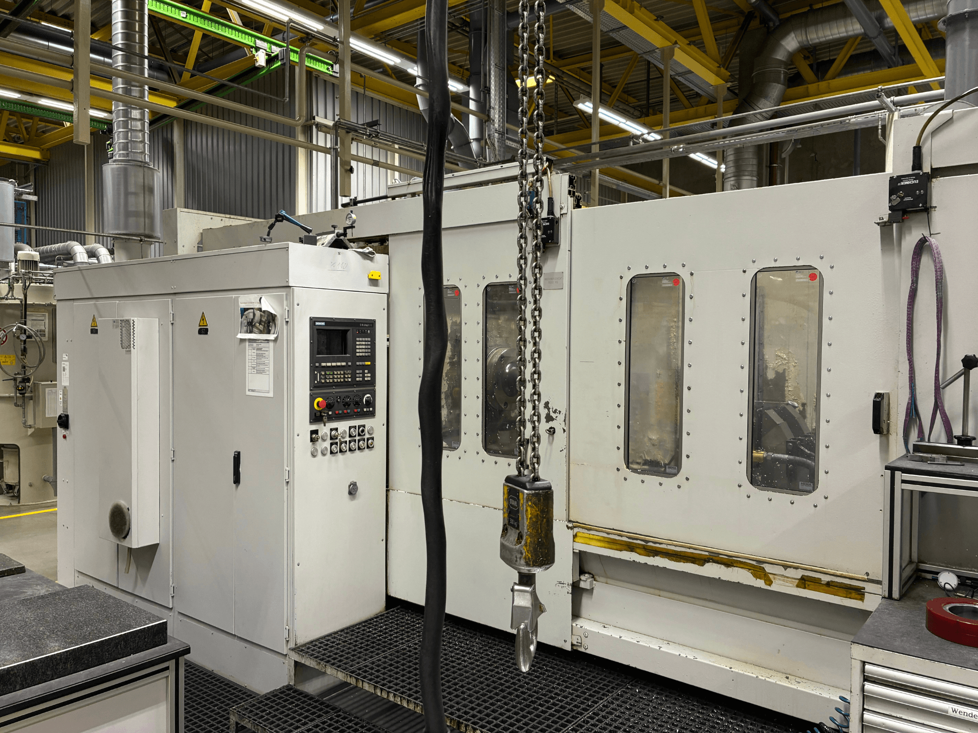 Máquina CNC industrial com painel de controlo, vista lateral; apresenta janelas, correntes e componentes metálicos num ambiente de fábrica.