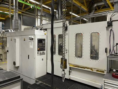 Máquina CNC industrial com painel de controlo, vista lateral; apresenta janelas, correntes e componentes metálicos num ambiente de fábrica.